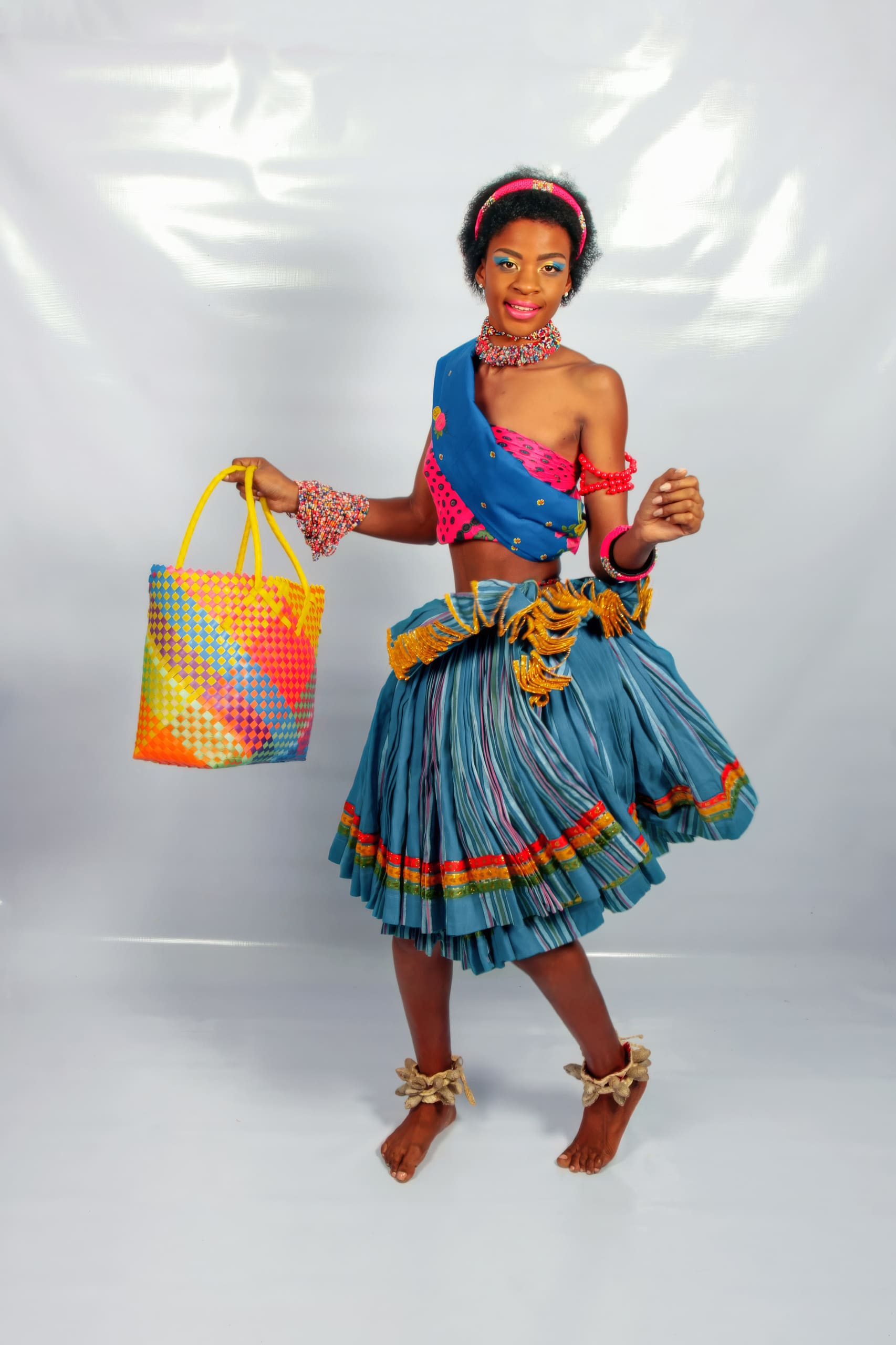 A smiling woman wears a colourful traditional outfit with a pleated blue skirt, red sash, bead jewellery, and anklets. She holds a large, colourful woven bag and stands barefoot against a plain white backdrop.