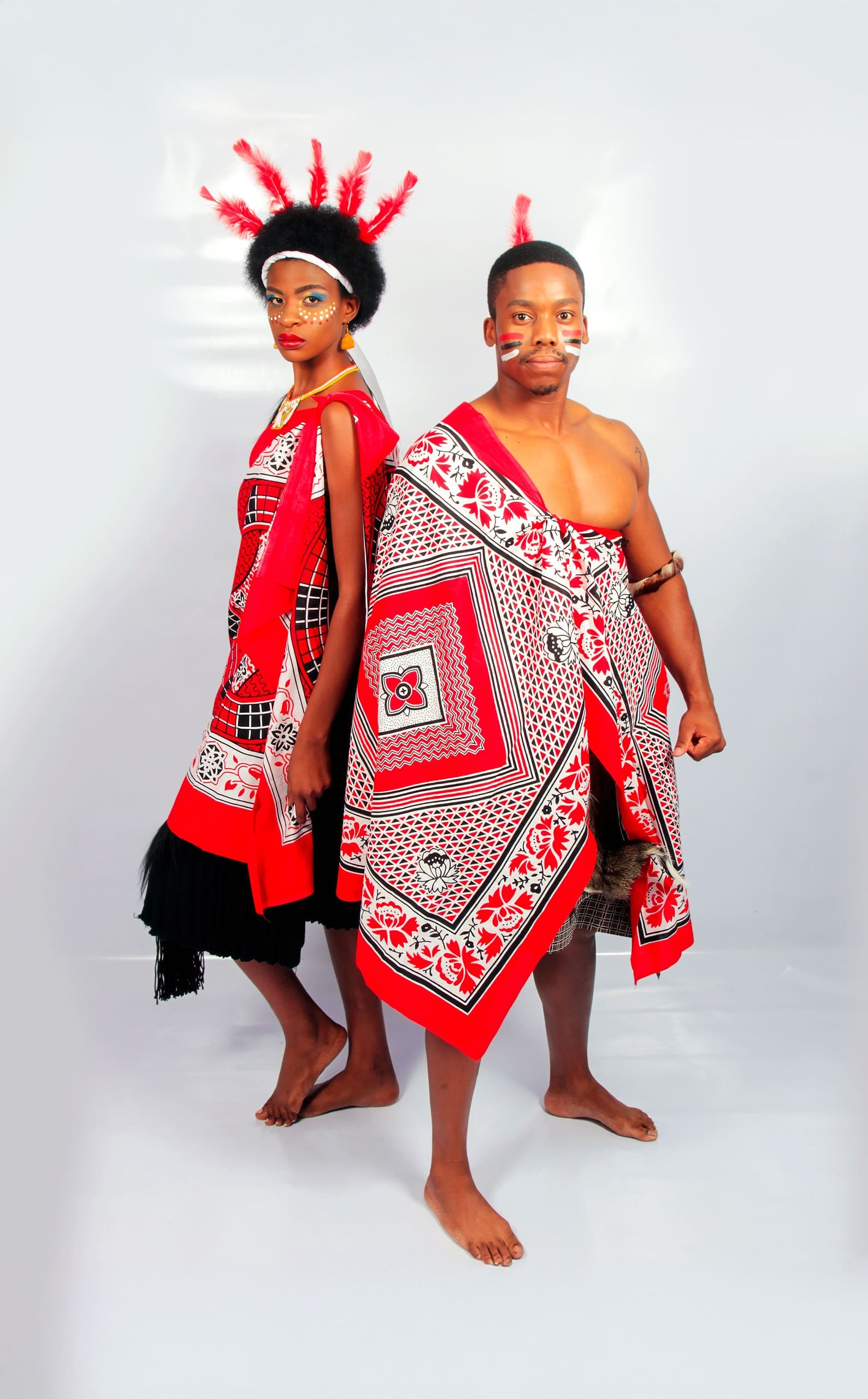 Two people wear vibrant red, black, and white traditional Swazi attire. The woman has face paint, a headdress with red feathers, and a patterned wrap. The man is bare-chested, with facial markings and a matching patterned cloth draped over his shoulder.