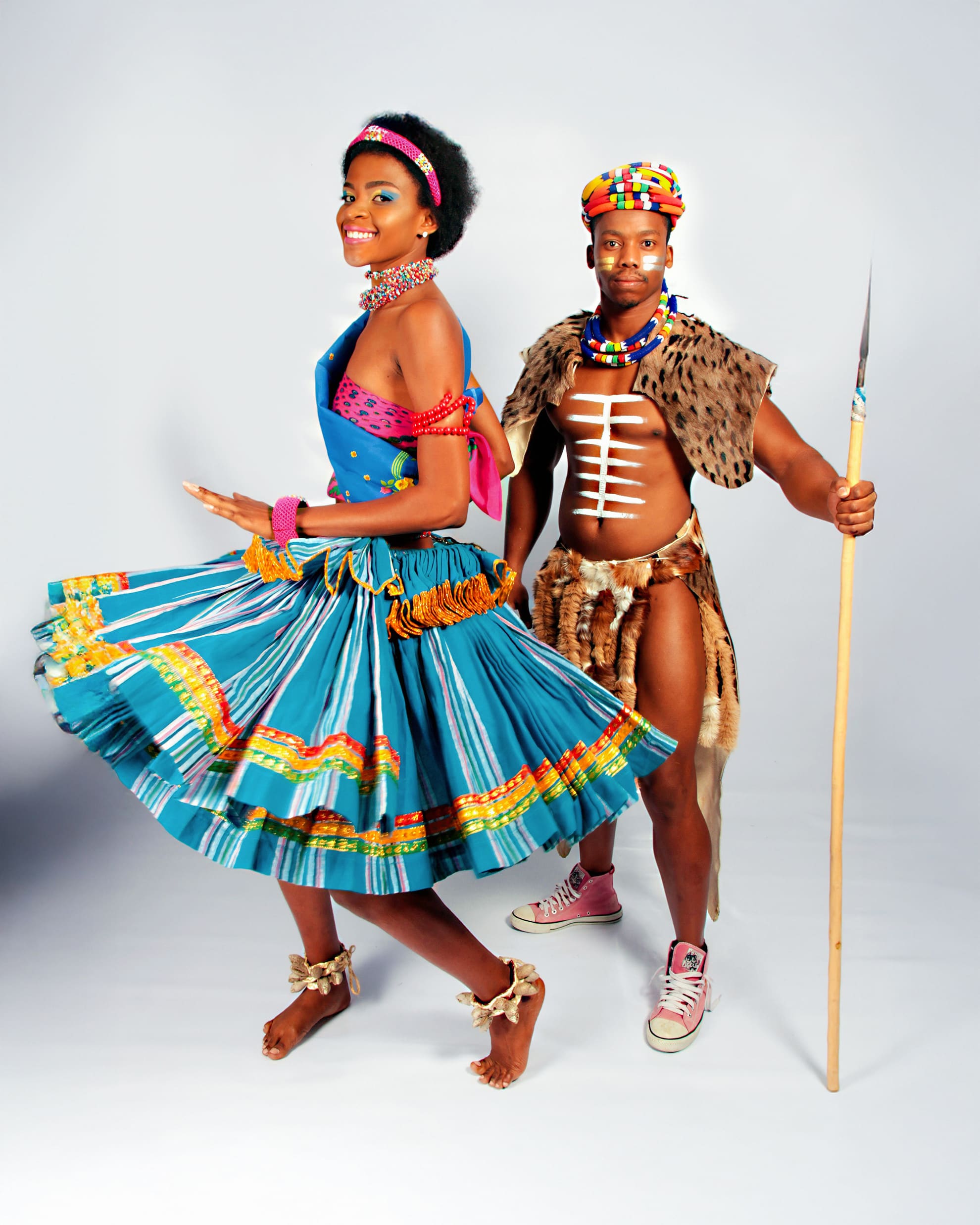 A woman and man in colourful traditional African clothing pose in a studio. The woman twirls in a blue patterned skirt with beaded jewellery, while the man wears animal print attire, beaded accessories, and holds a spear. Both are smiling against a plain background.