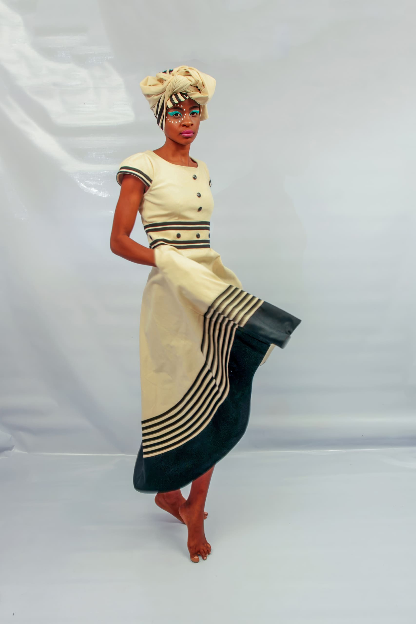 A person stands barefoot against a plain light backdrop, wearing a cream and black traditional dress with striped details. They have a matching headwrap and colourful face paint, looking directly at the camera whilst holding the skirt with one hand.