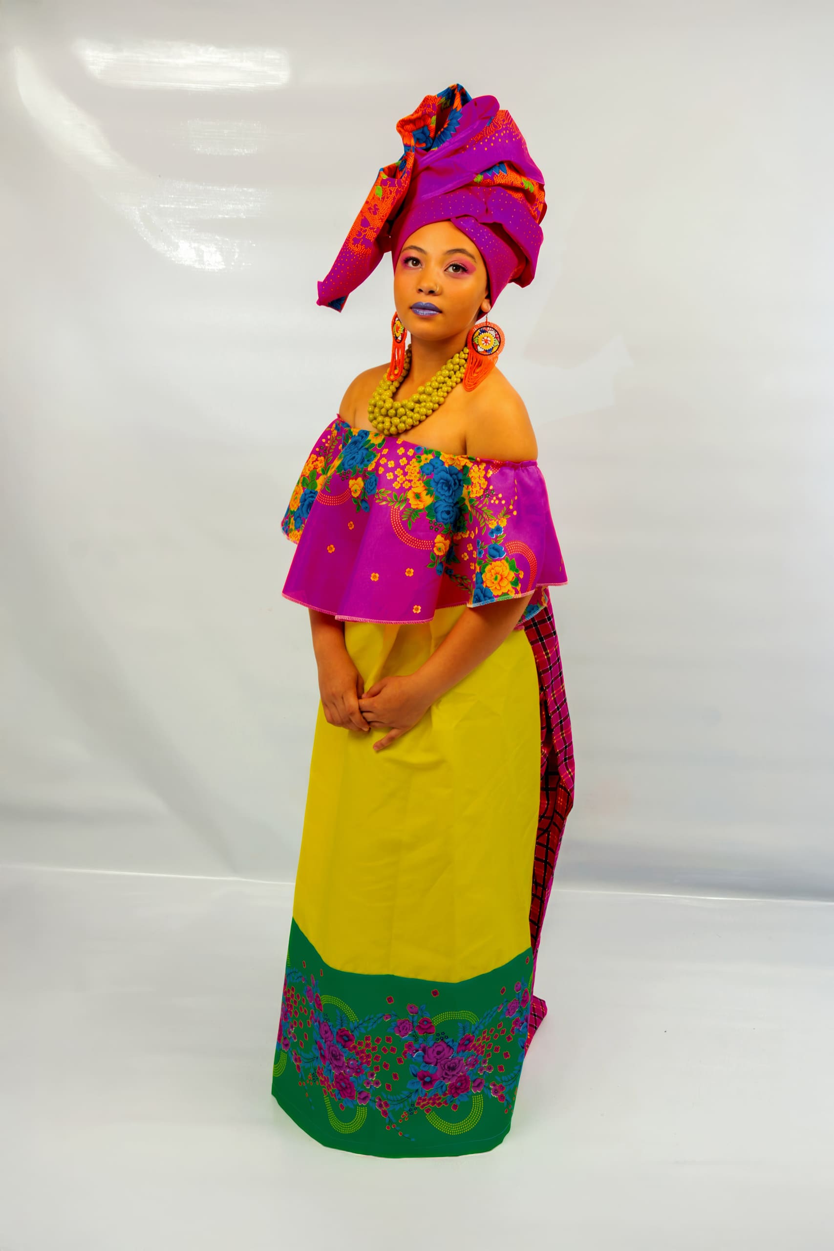 A woman stands against a plain white background, wearing colourful traditional African attire. She has a bright pink and yellow dress with green and floral patterns, a bold multi-coloured headwrap, large earrings, layered necklaces, and blue lipstick.