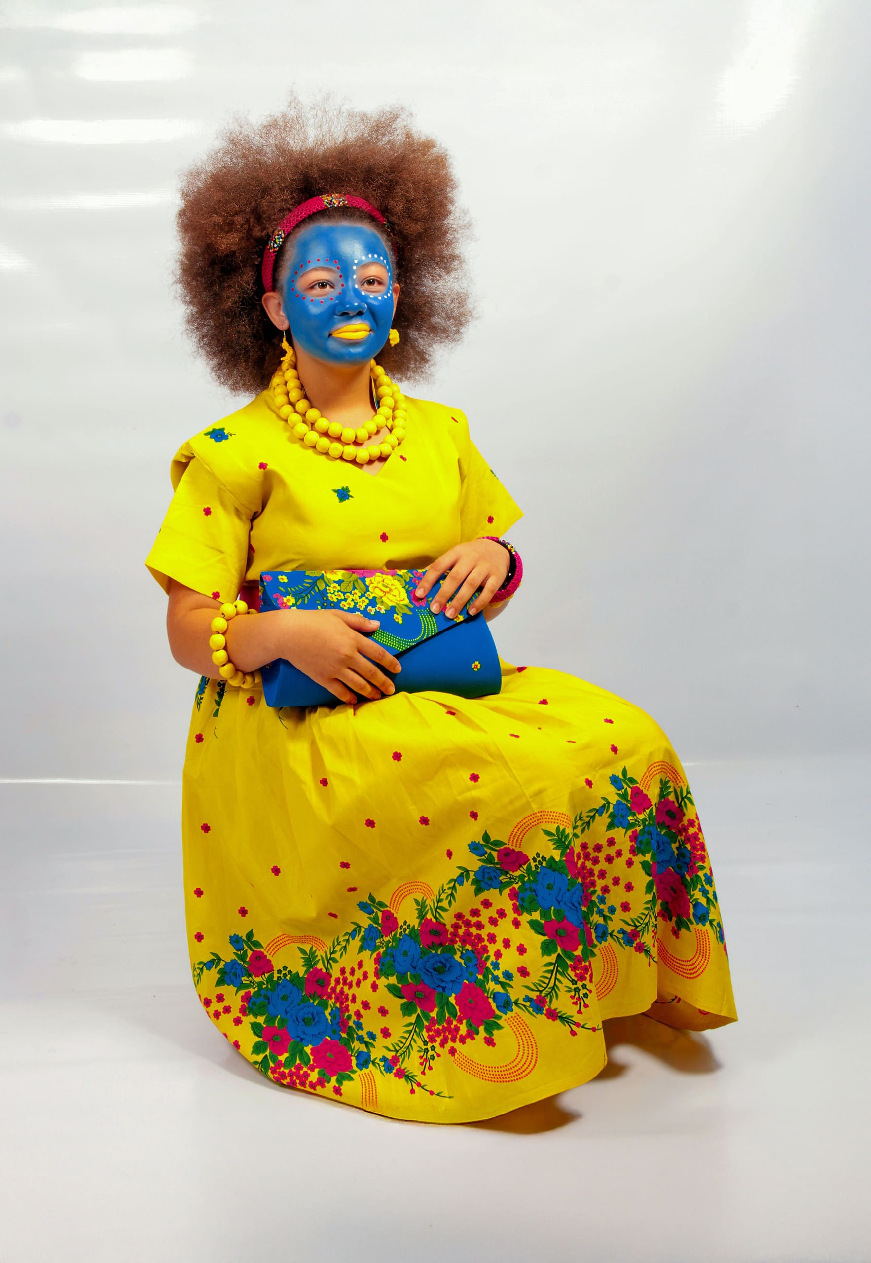 A woman with an afro hairstyle sits against a white backdrop, wearing a bright yellow floral dress, yellow jewellery, and holding a matching blue clutch. Her face is painted blue with white designs, and she wears a pink headband.