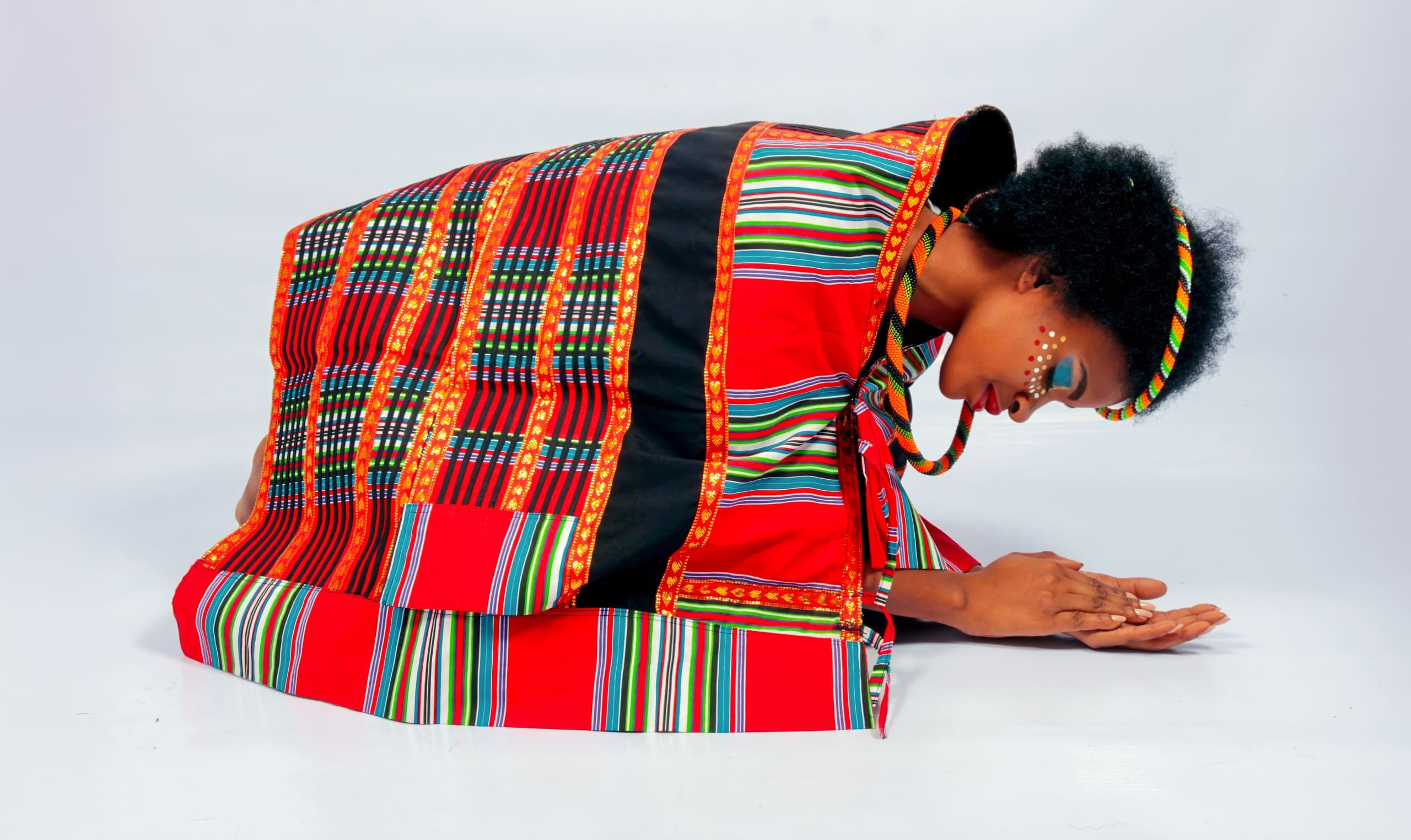 A person kneels and bows forward, touching their palms to the floor. They wear vibrant, patterned red and black traditional attire, with colourful beadwork and a beaded headband. Their face is painted with white dots and lines, against a plain light background.