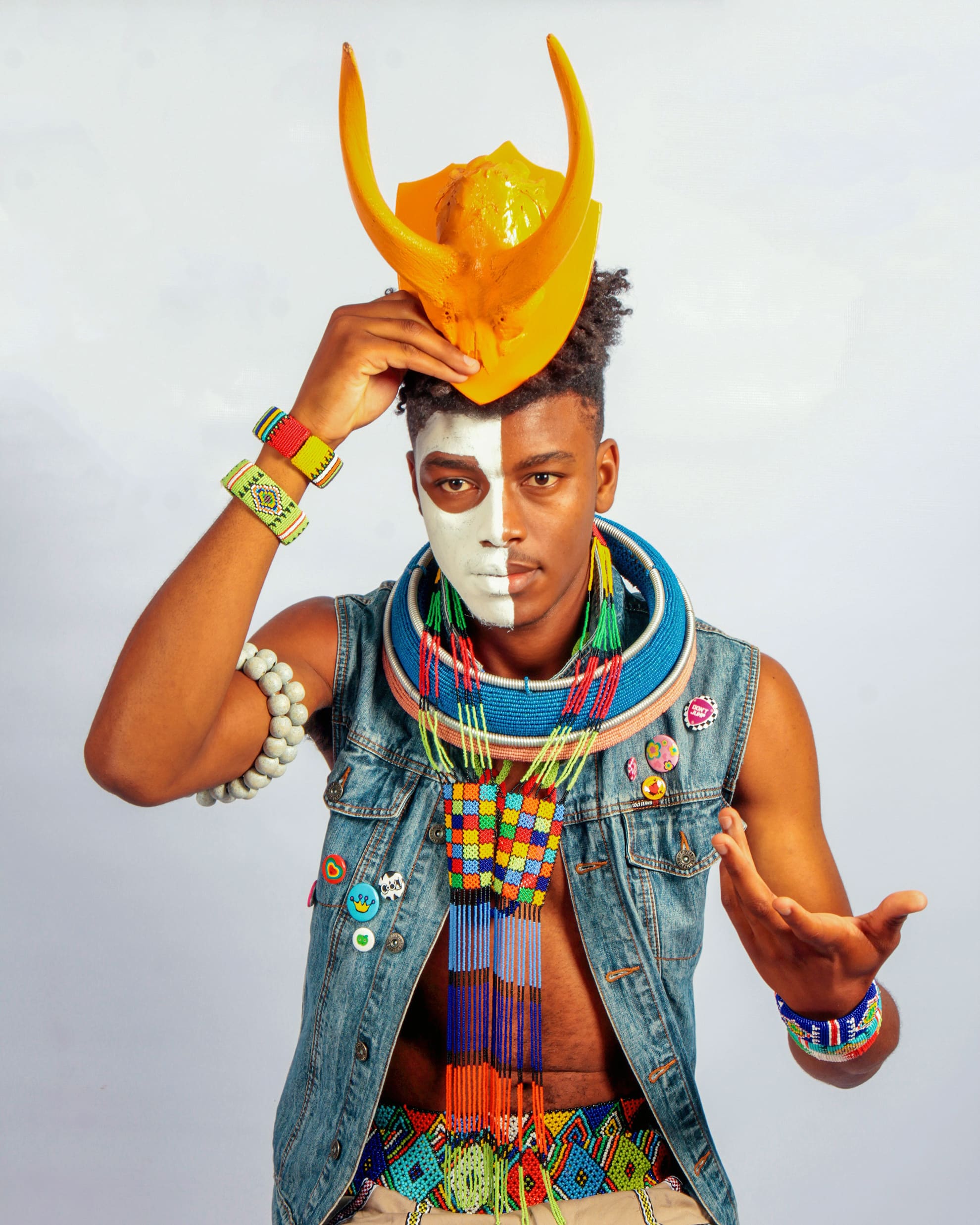 A person with half their face painted white wears colourful beaded jewellery, a sleeveless denim waistcoat, and patterned belts. They hold a yellow sculpted mask on their head, with their other hand raised. The background is plain and light-coloured.