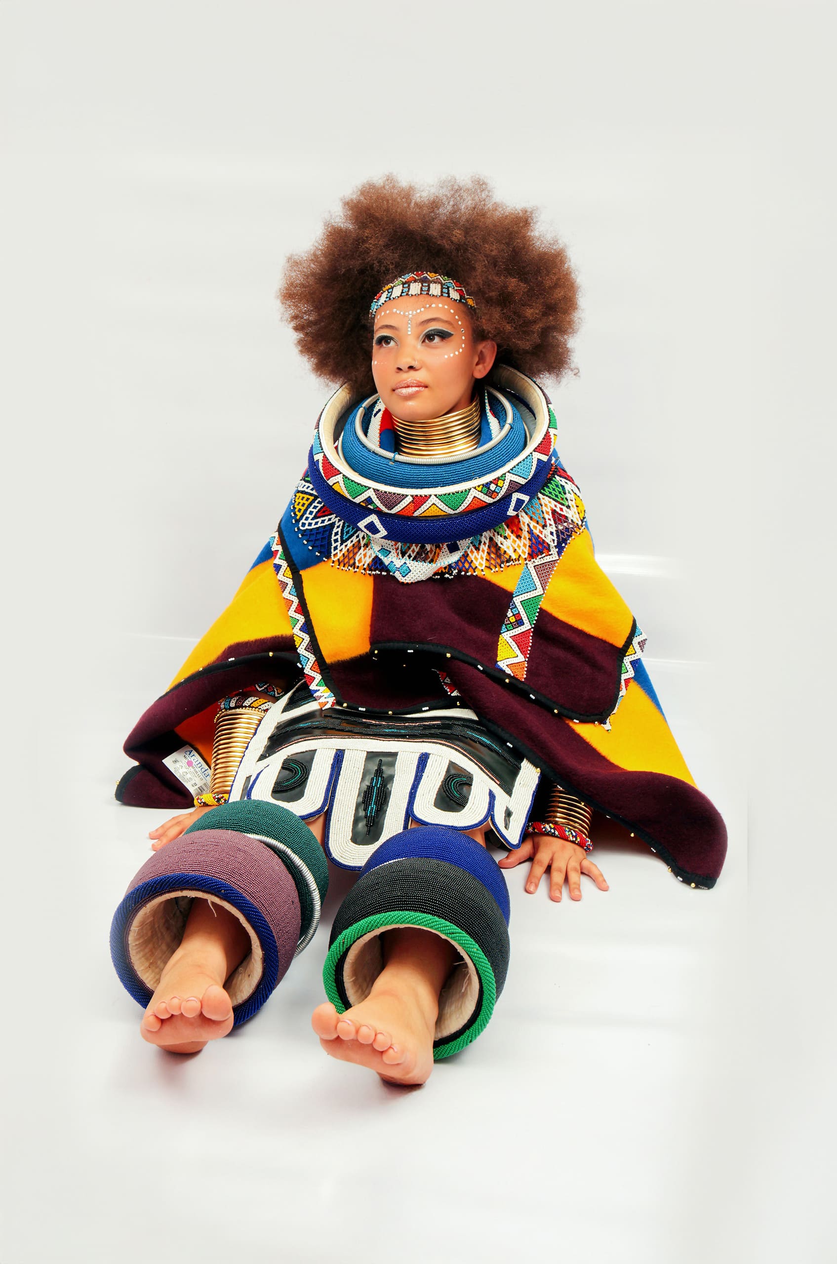 A woman with an afro hairstyle sits barefoot on a white background, wearing vibrant, geometric, multicoloured Ndebele attire with beaded necklaces, a headband, arm and leg rings, and a patterned cape. Her expression is calm and dignified.