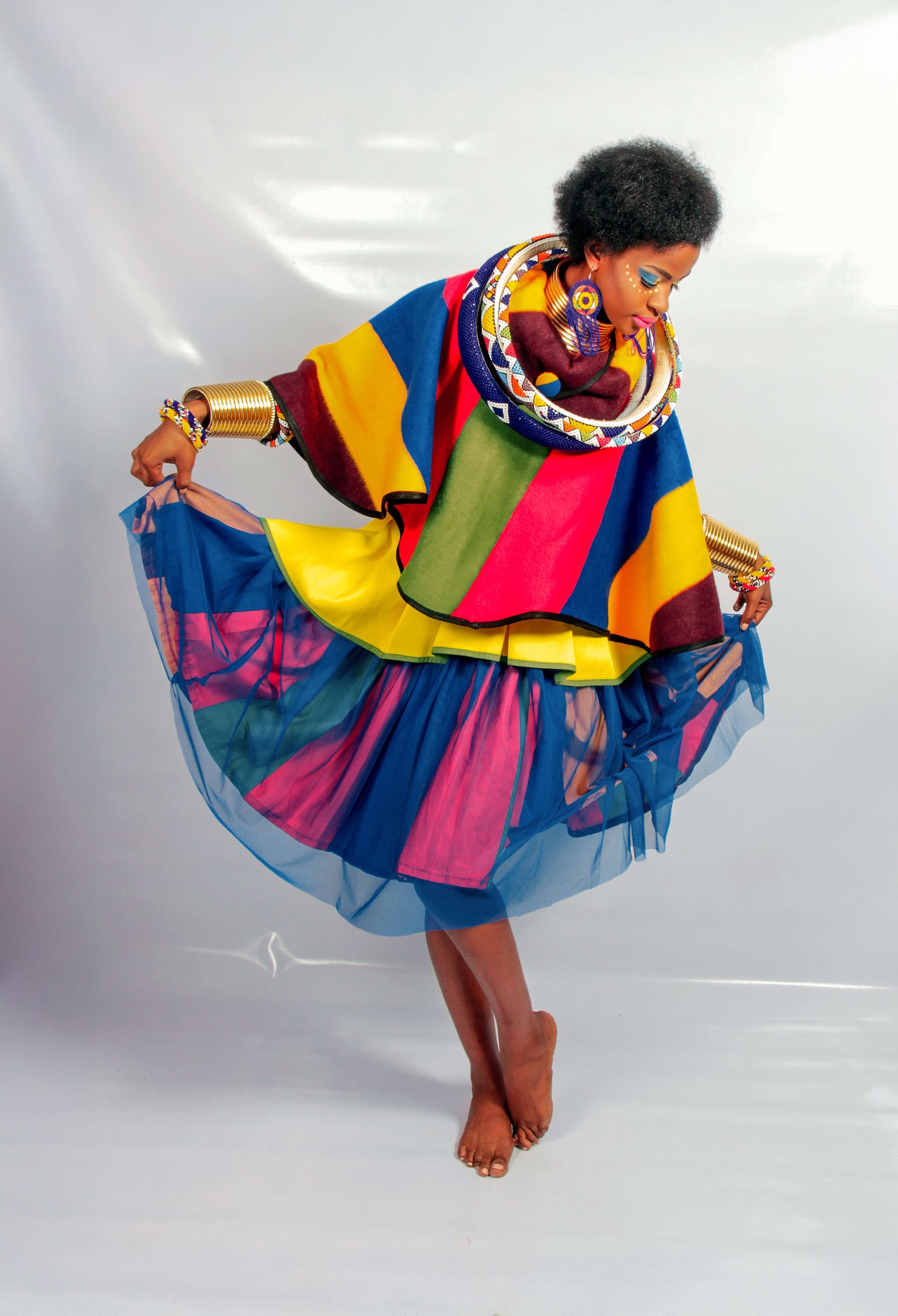 A barefoot woman poses gracefully in a multicoloured outfit with bold yellow, red, blue, and green stripes. She wears gold bangles, colourful necklaces, and a sheer blue skirt layered over a bright underskirt. Her makeup is vibrant and artistic.