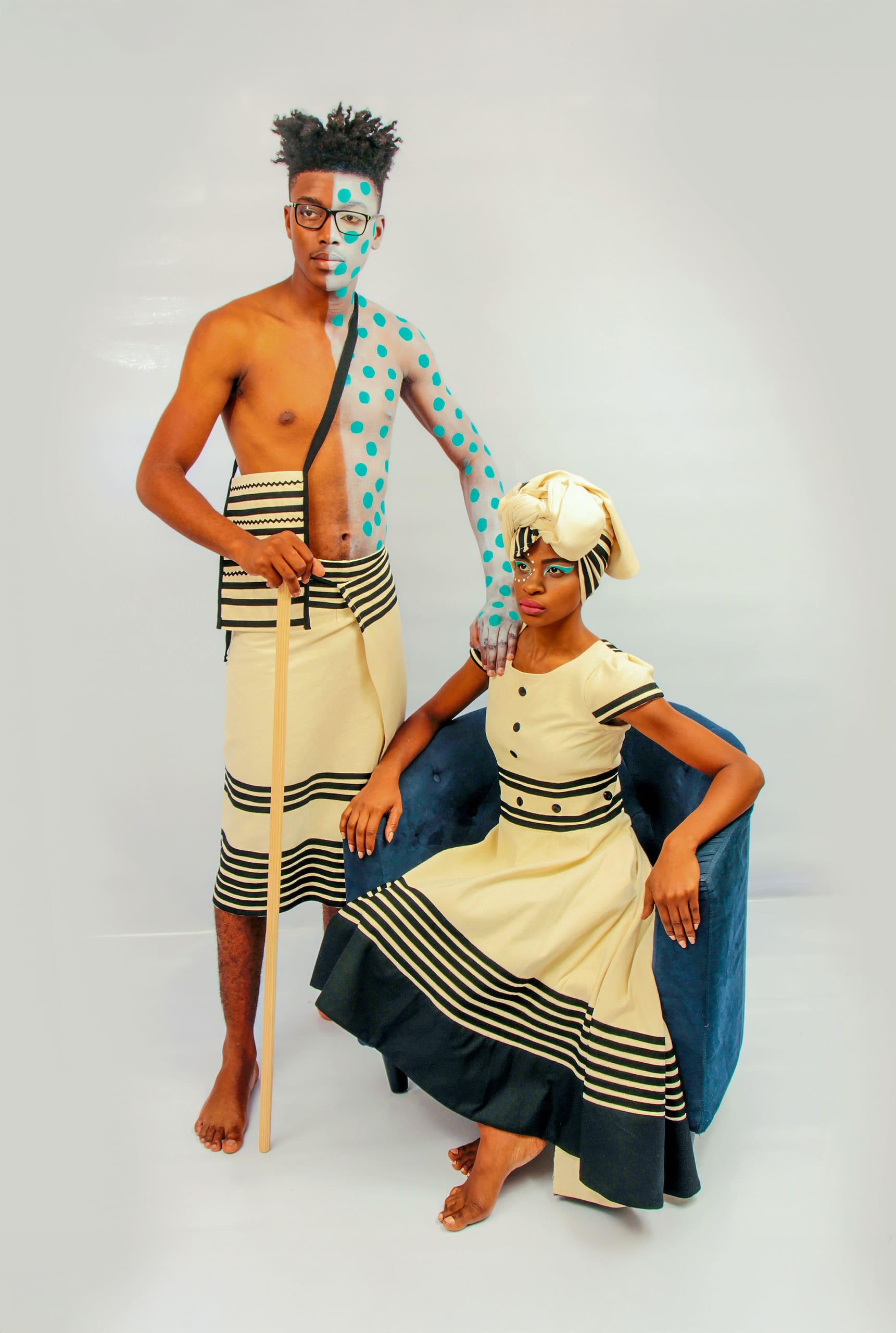 A barefoot man and woman pose in matching beige and black traditional attire. The man stands holding a wooden staff, his face and torso painted with turquoise dots and lines. The woman sits on a blue chair with a matching headwrap and face paint, both looking serious.