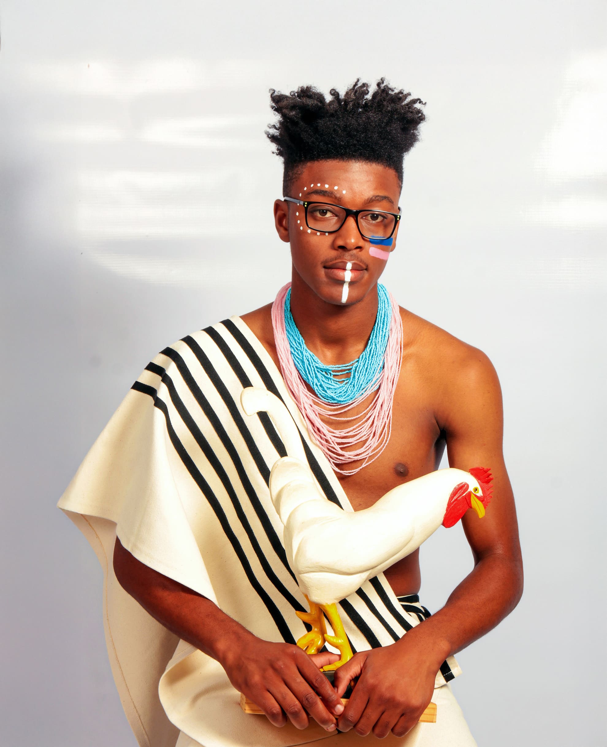 A young man wears beaded necklaces, traditional attire with black and white stripes, and face paint. He has short, styled hair and glasses, and sits holding a white cockerel figurine with a red comb and yellow beak, against a plain light background.