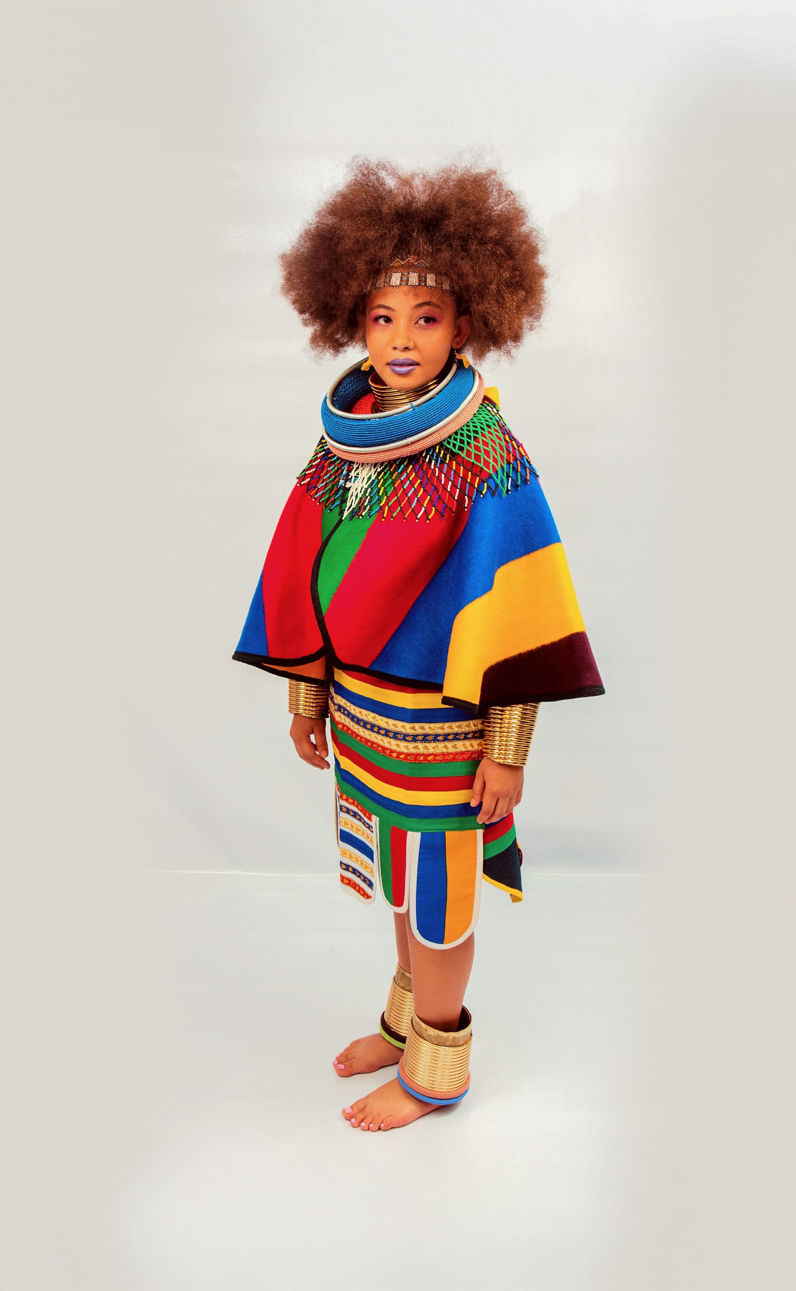 A young child with a large afro hairstyle stands barefoot, wearing vibrant, traditional African attire in red, blue, green, and yellow, with beaded jewellery, a patterned wrap, and decorative anklets, set against a plain light background.