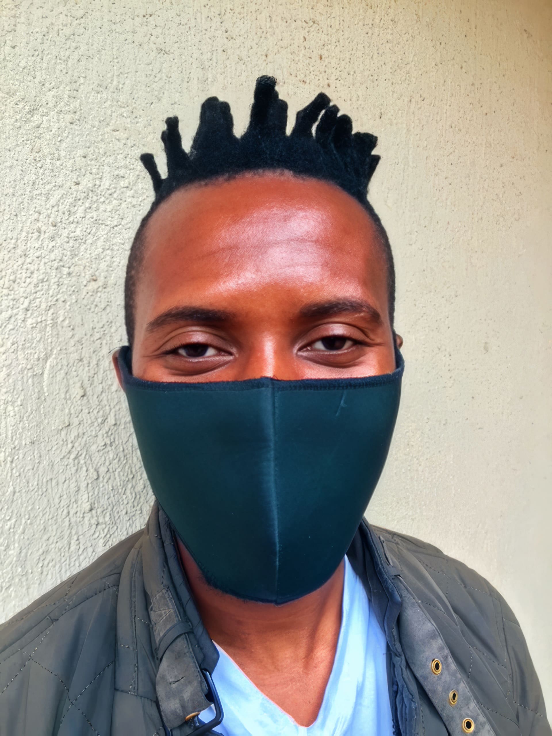 A person with short, styled black hair is wearing a dark face mask and a green quilted jacket over a light blue shirt, standing in front of a textured beige wall. Only their eyes, forehead, and upper cheeks are visible.