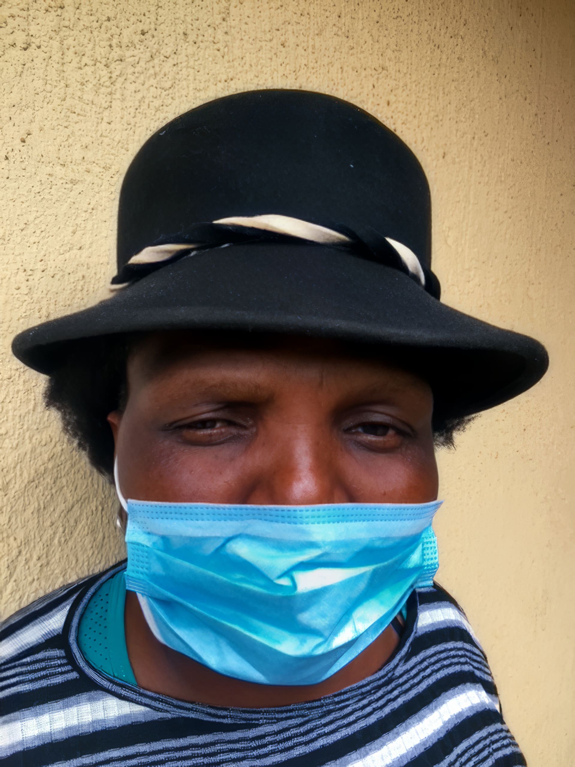 A person wearing a black hat with a white-and-black band and a striped shirt is facing the camera. They have a blue surgical mask covering their nose and mouth. The background is a beige textured wall.