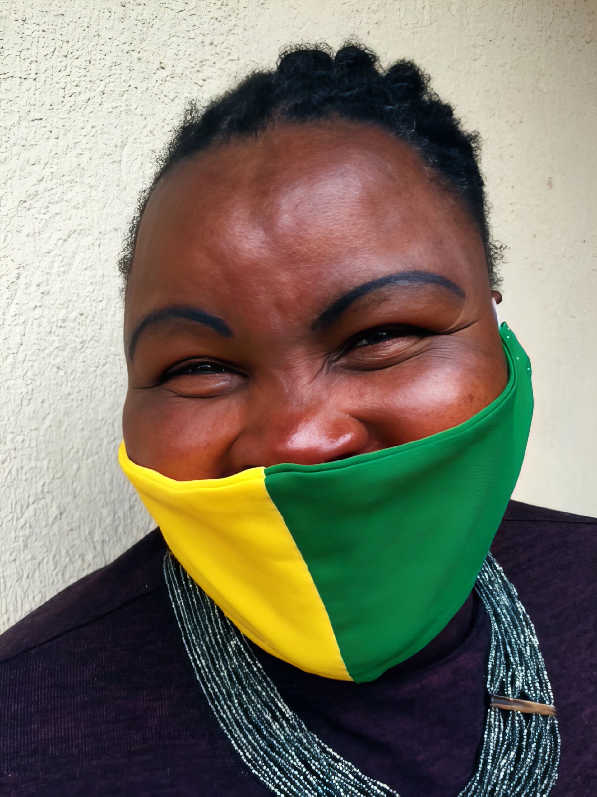 A smiling person with plaited hair wears a half-yellow, half-green mask pulled below their nose, revealing their wide smile. They have prominent, arched eyebrows and a layered beaded necklace, standing against a textured beige wall.