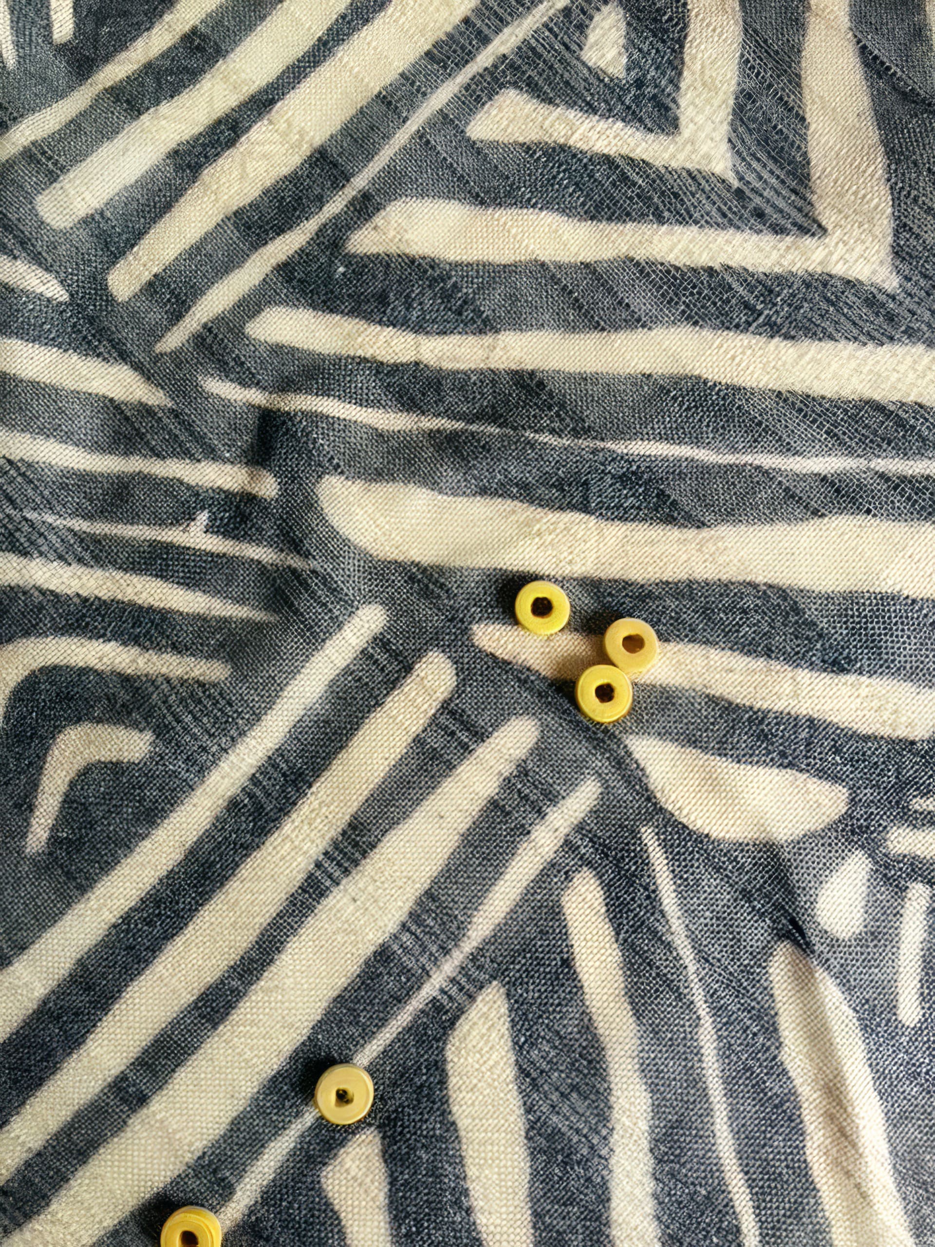 A close-up of a fabric with white and dark blue geometric, zigzag patterns. Scattered on the fabric are six small, round, yellow beads. The surface shows a textured, woven appearance with visible threads and folds.