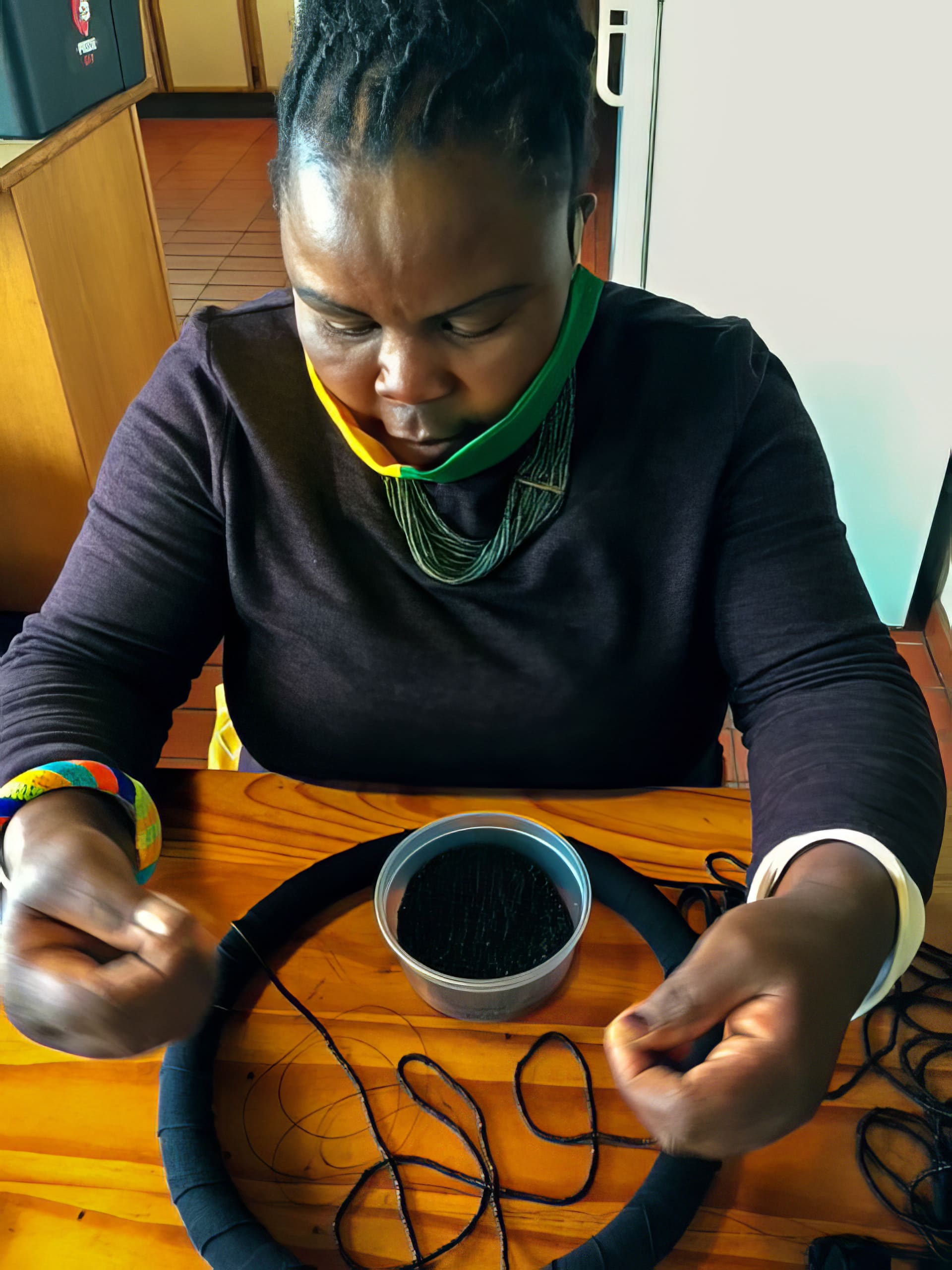 A woman with plaited hair, wearing a black top and colourful beaded bracelet, sits at a wooden table, threading black beads onto a circular frame. A tin of black beads is in front of her, and she wears a green and yellow mask below her chin.