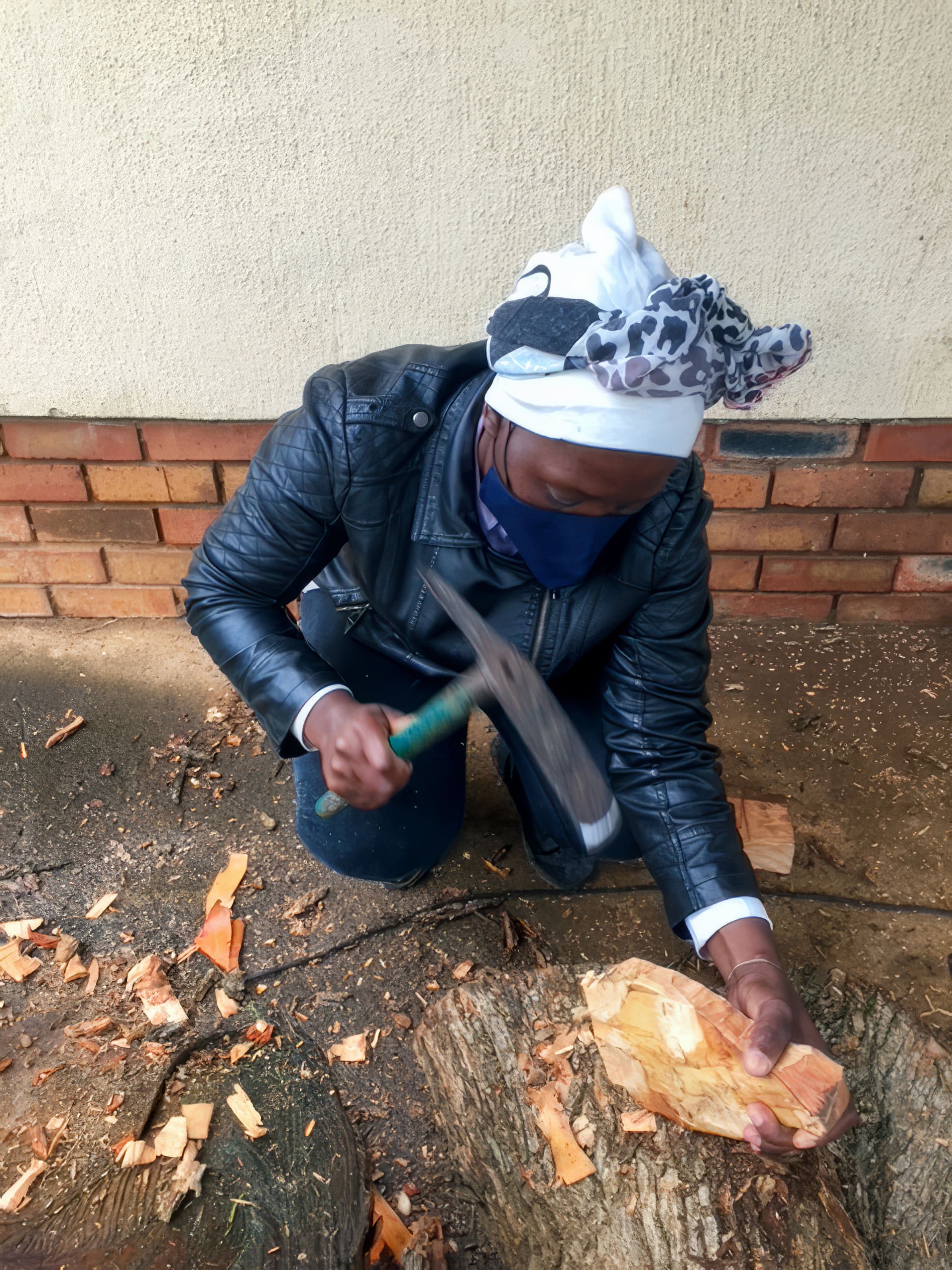 A person wearing a black jacket, blue face mask, and white headscarf kneels on the ground, using a hammer on a piece of wood. Wood shavings scatter around. The background features a brick and plastered wall.