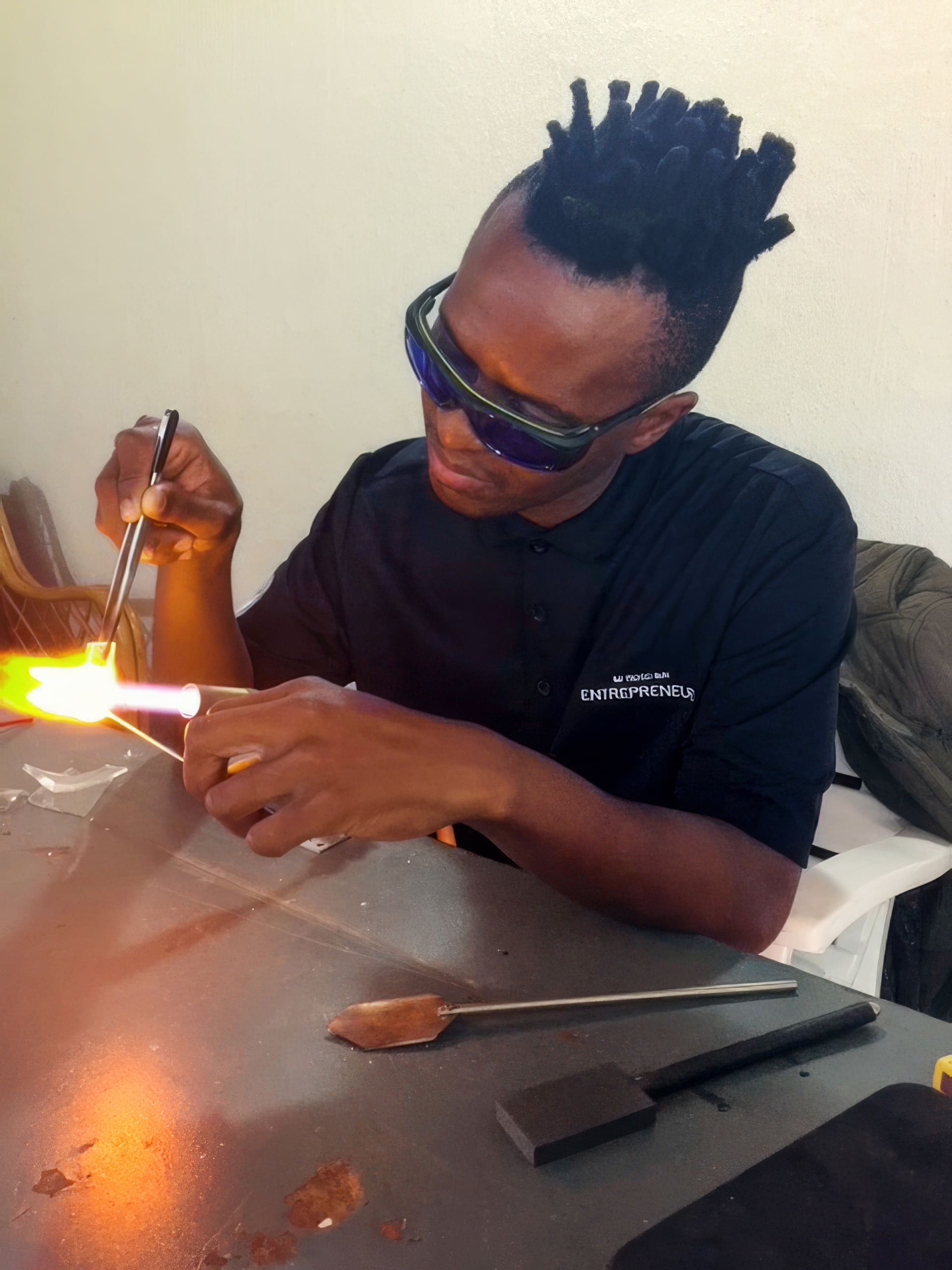 A person with short, spiked hair and protective glasses shapes molten glass with tools and a torch at a worktable. They wear a black shirt reading 