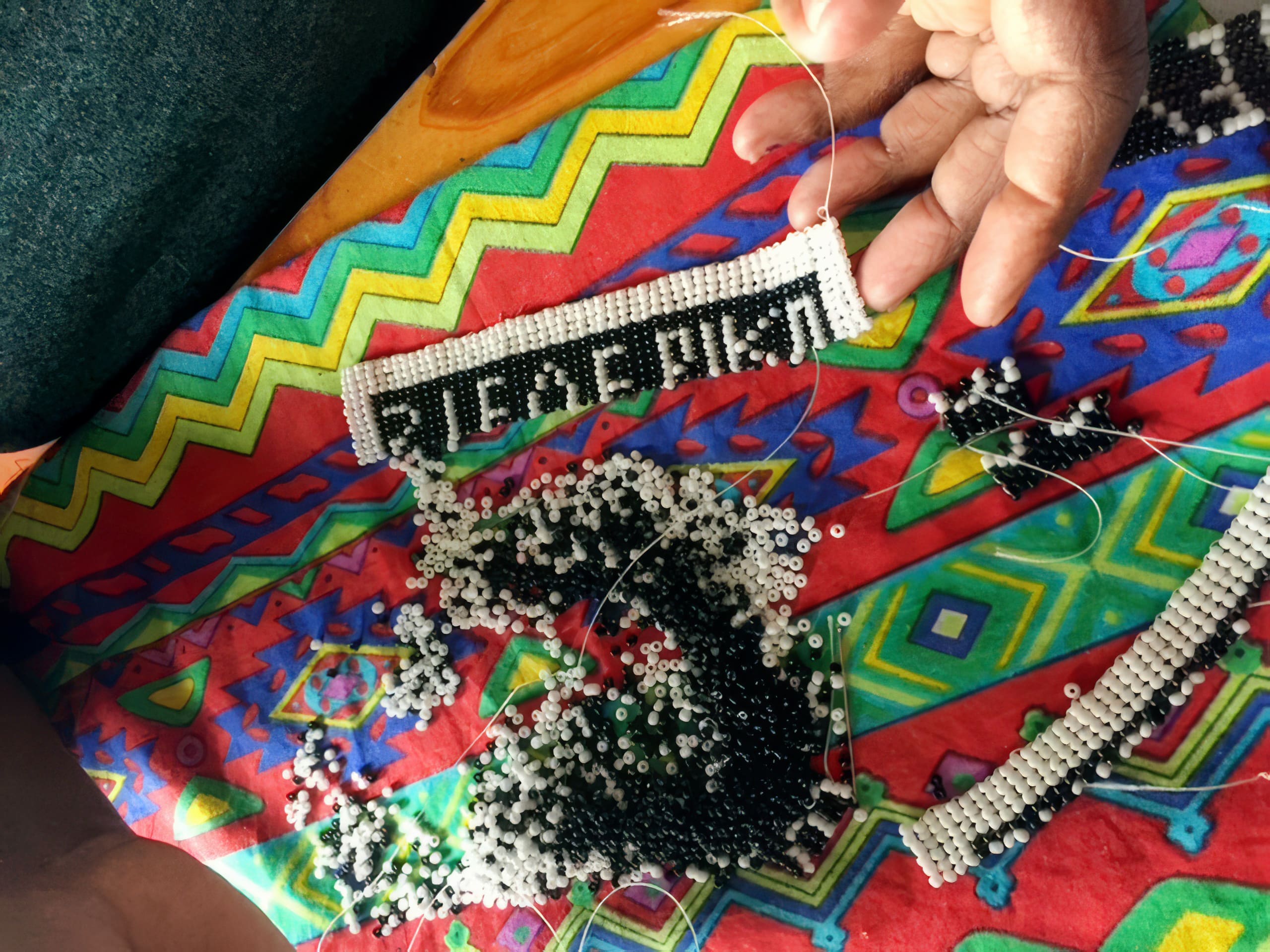 A hand holds a beaded craft spelling “AFRICA” in black and white beads on a colourful fabric with geometric patterns. Loose black and white beads and partially finished beadwork are scattered on the bright, patterned surface.