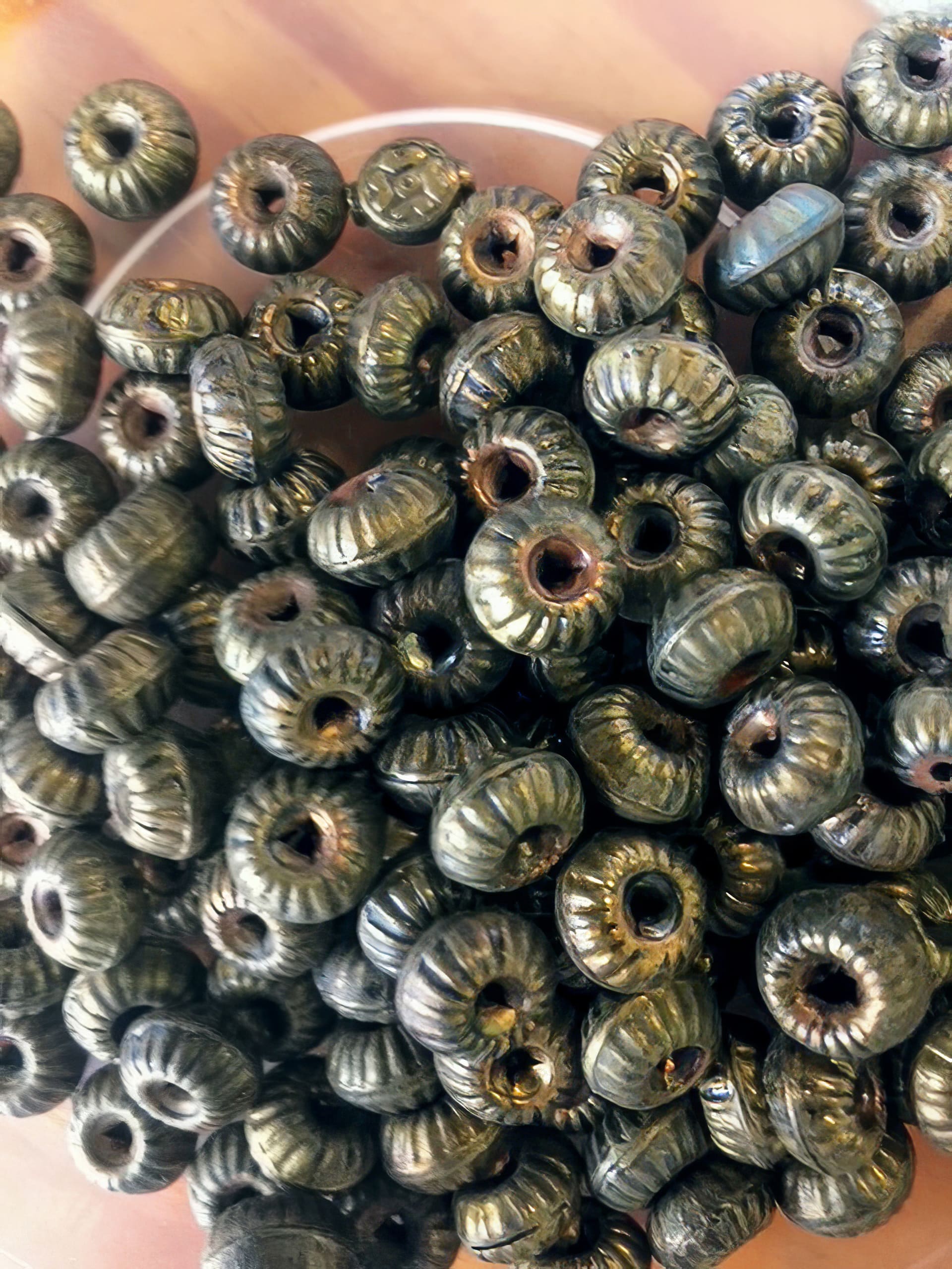 A close-up photo of many round, dark green glass beads with a metallic sheen and grooved, ridged surfaces. Each bead has a central hole, and the beads are piled together in a bowl, catching the light to show their textures.