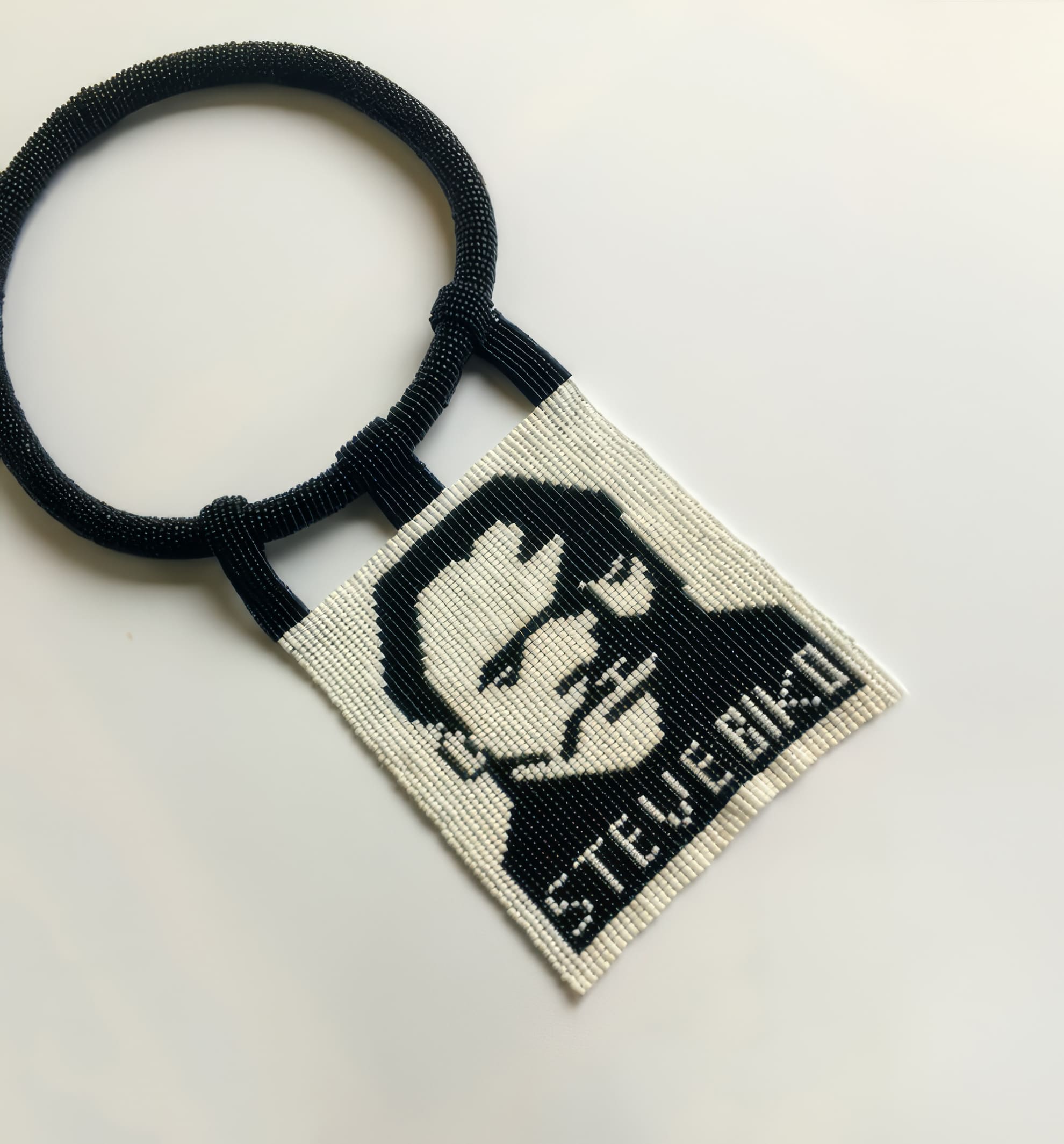 A beaded necklace with a rectangular pendant in black and white, featuring a stylised portrait of a man’s face and the text “STEVENBAD” below. The pendant is attached to a thick, round beaded collar by two narrow beaded straps.