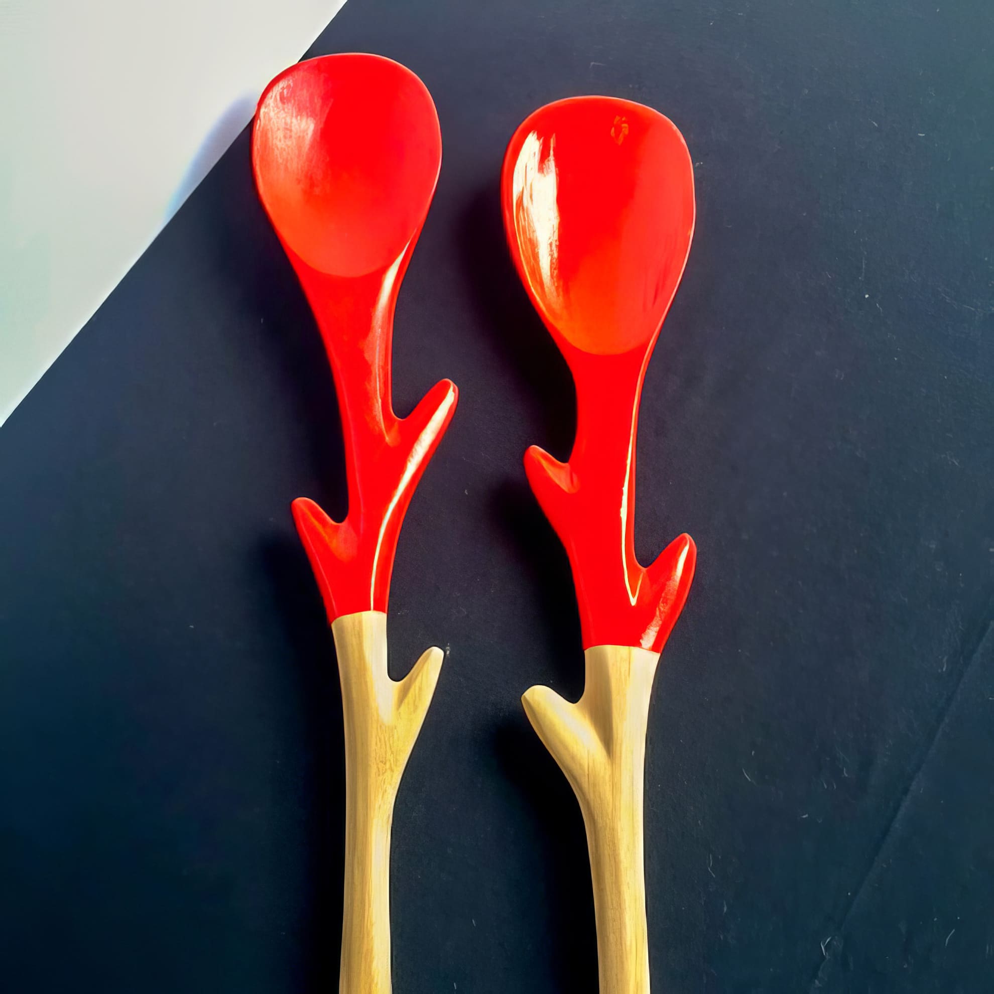 Two wooden spoons with red tops and handles shaped like tree branches are placed side by side on a black and white background, creating a vibrant and artistic contrast.