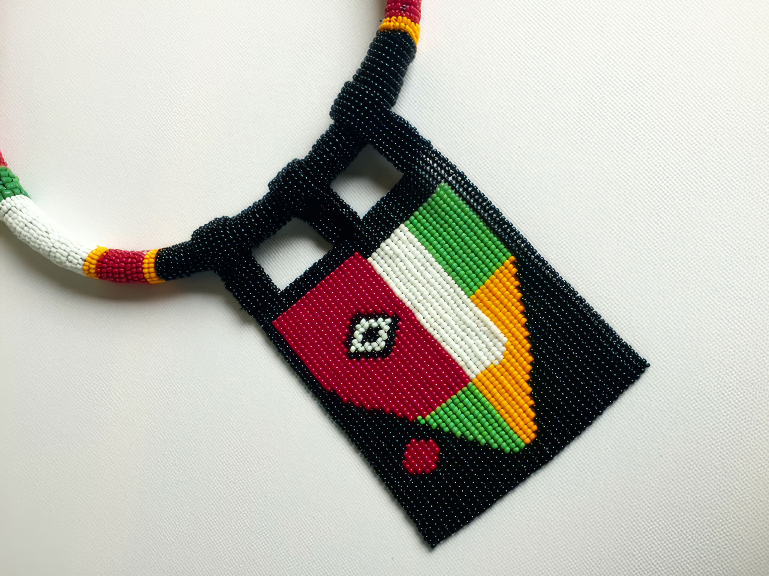 A handcrafted beaded necklace with a geometric pendant. The pendant features blocks of red, green, white, yellow, and black beads, and a diamond pattern in the centre. The beaded cord is multicoloured, including red, yellow, white, green, and black.