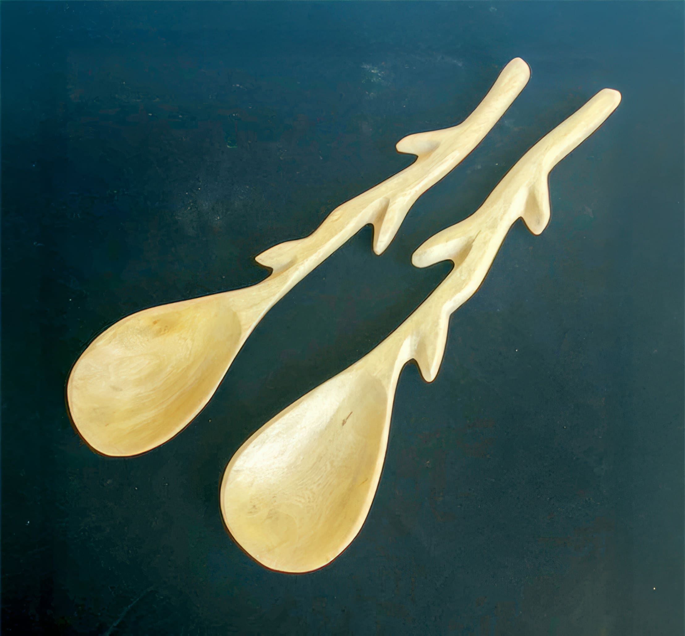 Two wooden spoons with handles shaped like tree branches, featuring multiple offshoots, are placed side by side on a dark, solid background. The spoons have a smooth, polished texture and a light natural wood colour.