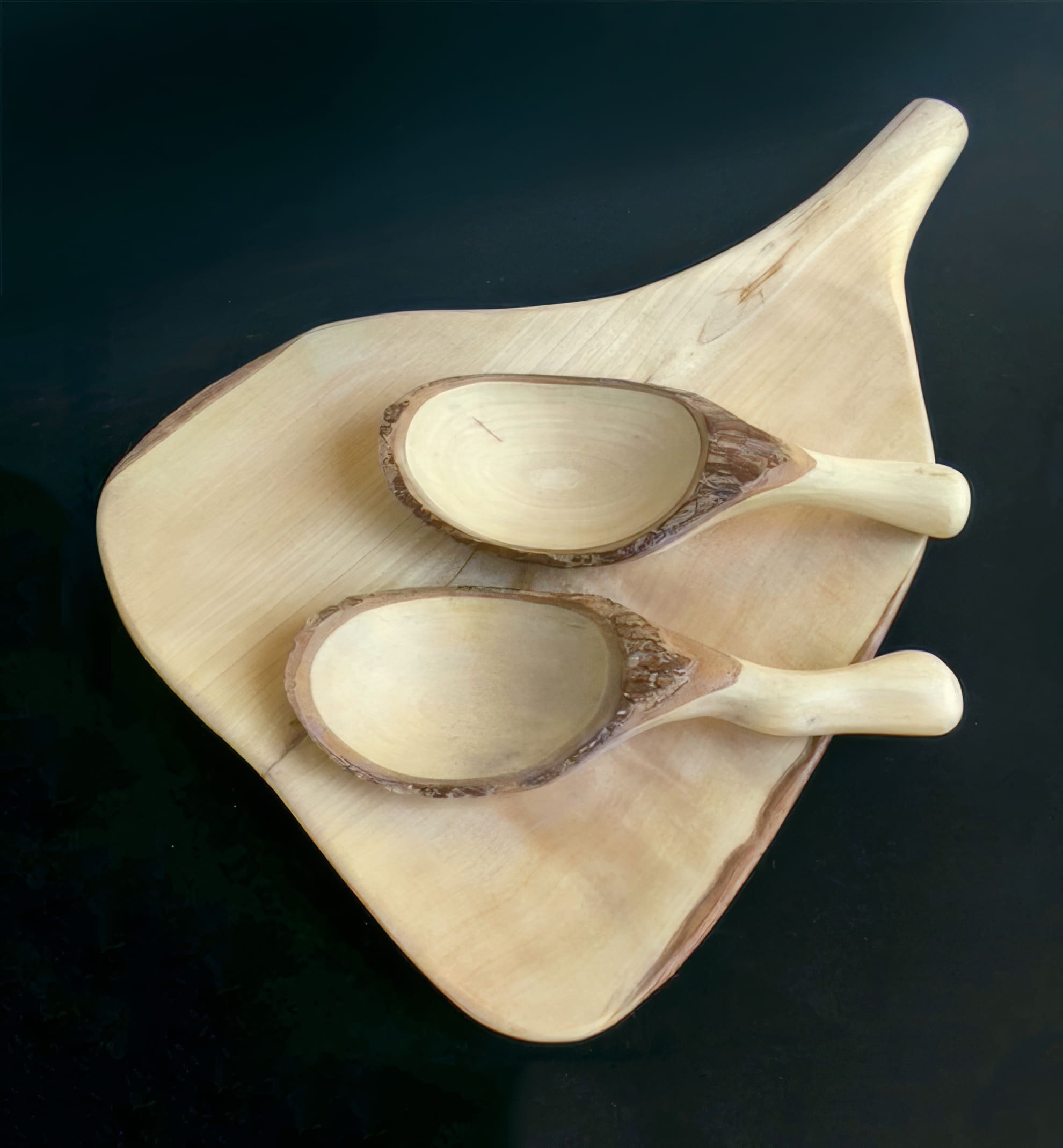 Two hand-carved wooden spoons with bark edges rest on a light wooden chopping board shaped like a leaf, all set against a dark background. The spoons and board have a natural, rustic appearance with smooth finishes.