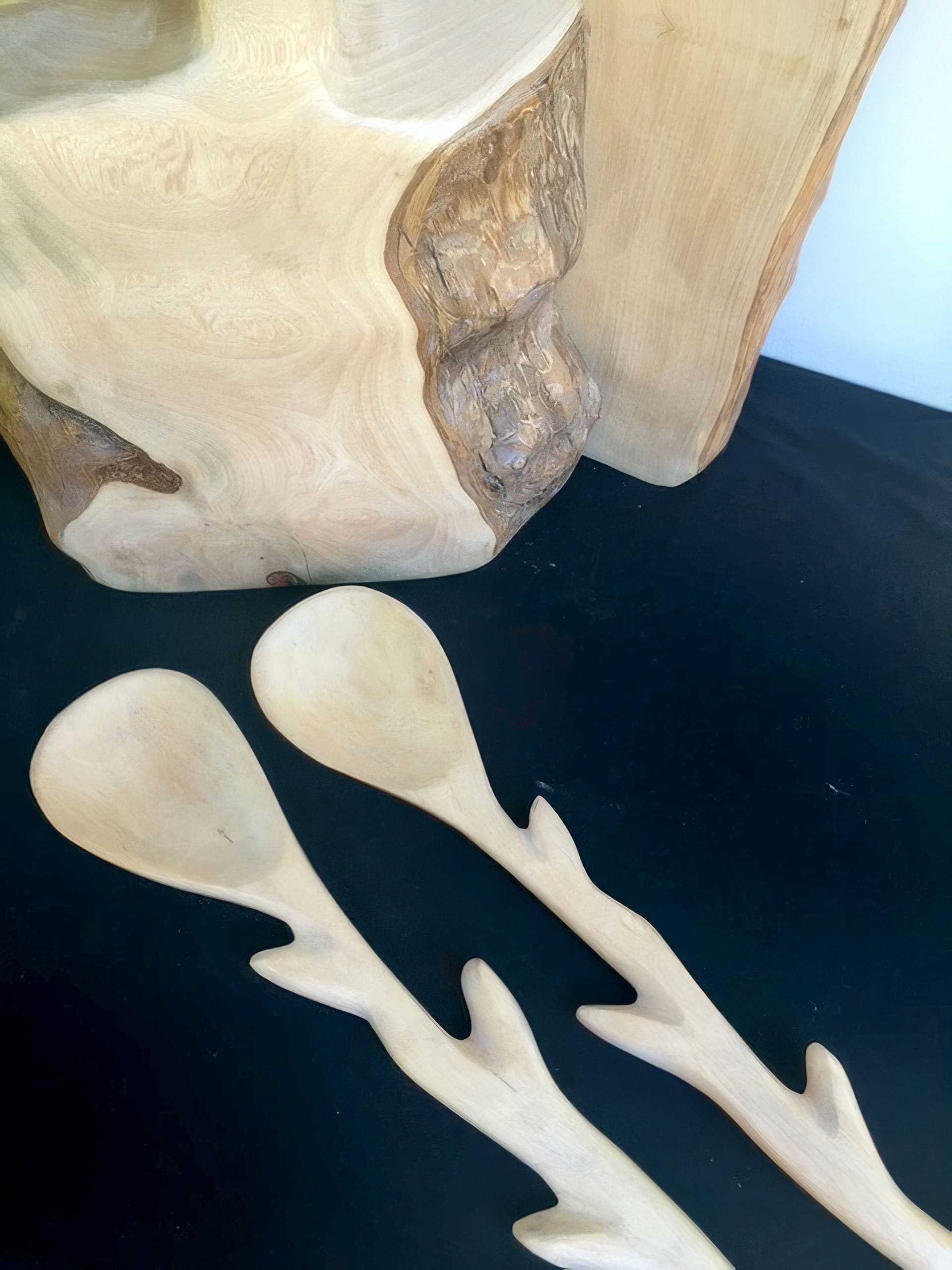 Two light-coloured wooden salad servers with branch-like handles lie on a black surface. Behind them is a large wooden chopping board featuring a natural bark edge and smooth, polished grain patterns. The scene highlights the wood's organic textures.