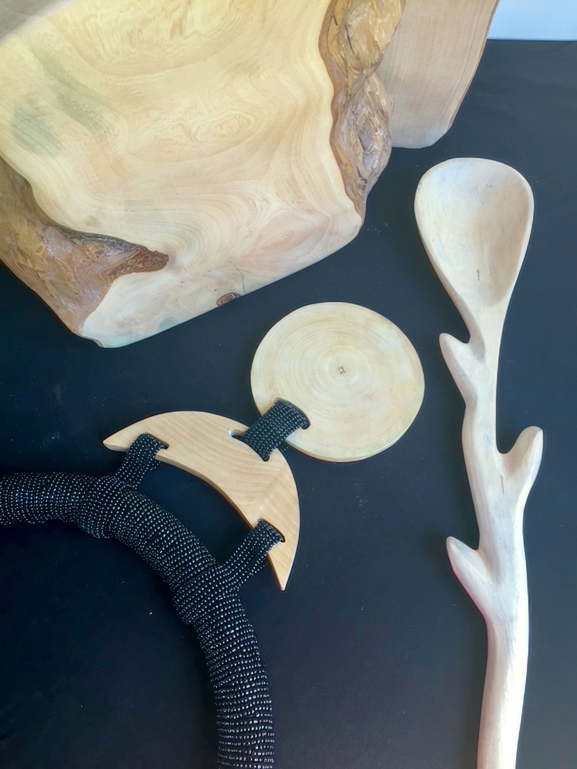 A photo shows a set of hand-carved wooden objects on a black surface: a large, blocky wood piece, a smooth circular disc, a crescent-shaped item with black rope, and a spoon with a handle shaped like a tree branch.