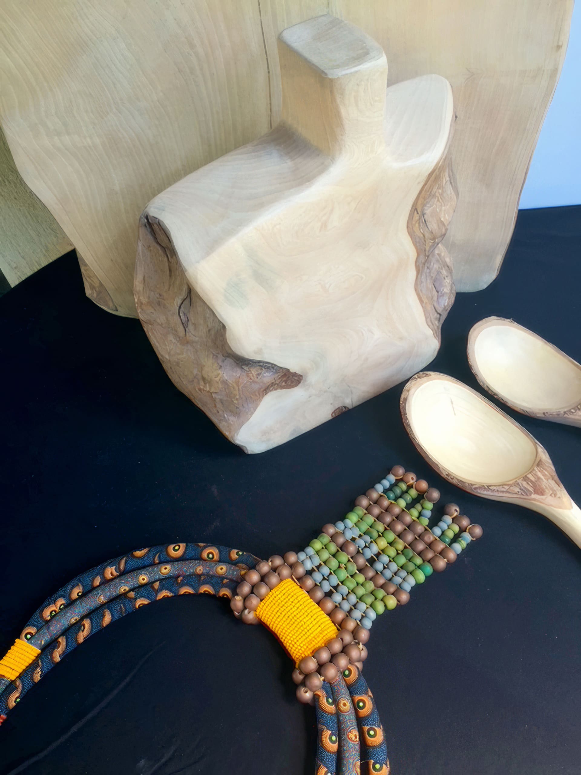 A hand-crafted wooden bust and two light wooden spoons are displayed on a black surface. In front of them lies a beaded necklace with green, brown, and yellow beads, and patterned black cords. The background is neutral and uncluttered.