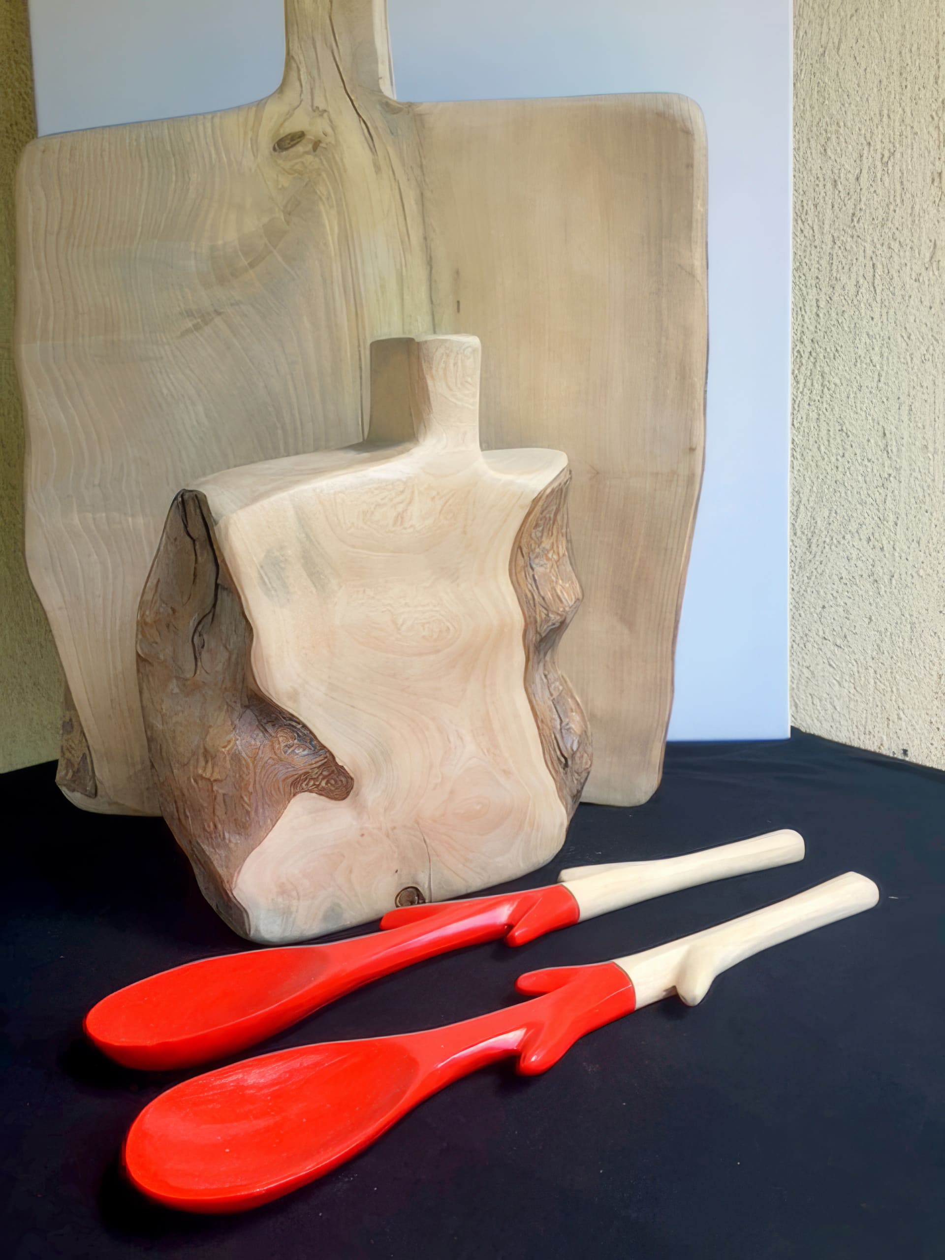Two large wooden chopping boards with natural edges stand upright on a black surface. In front, two wooden spoons with bright red handles and tips rest side by side. The background is neutral, with soft lighting highlighting the wood grain.