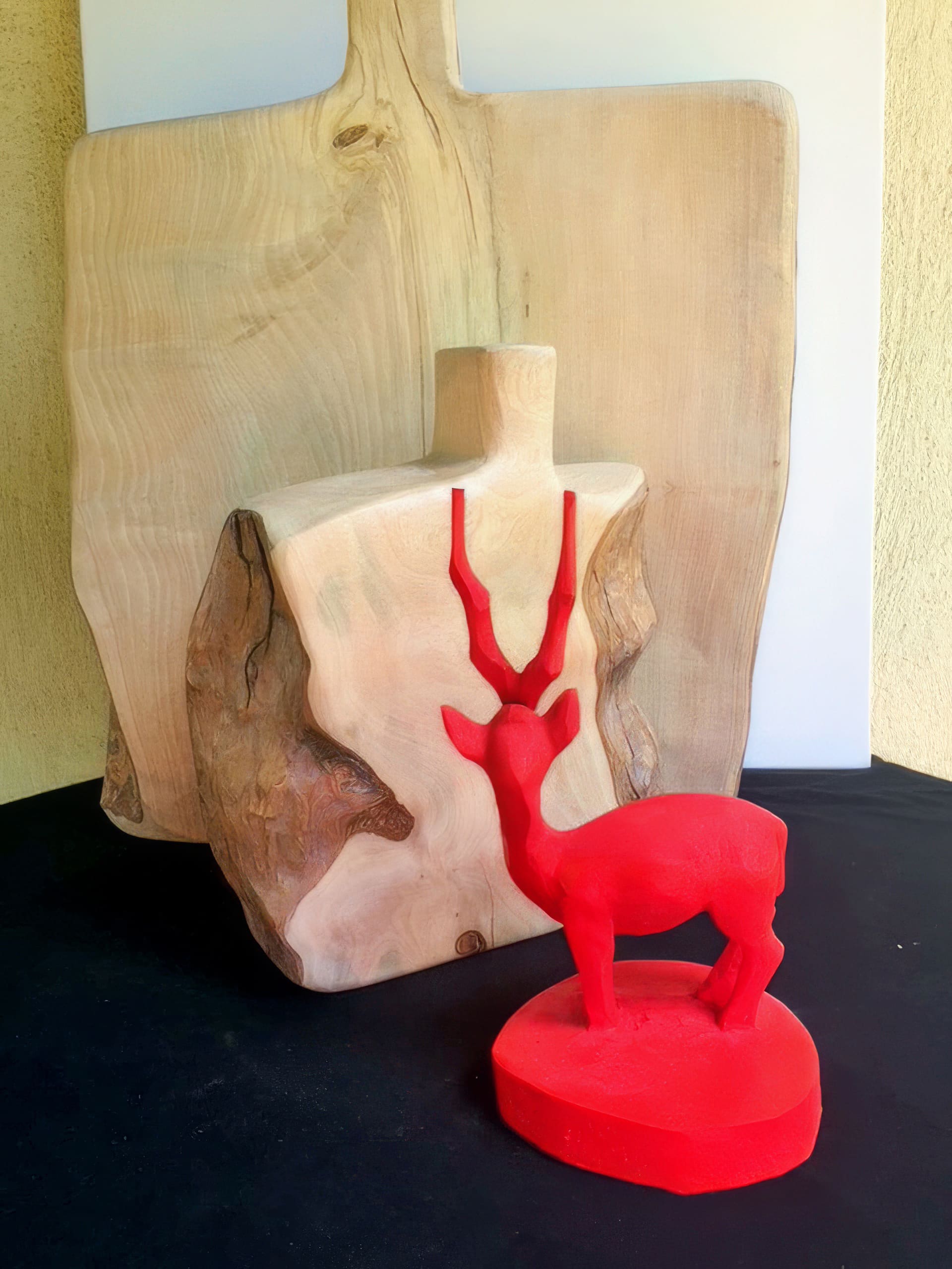 A red sculpture of an antelope stands on a red base in front of two large wooden chopping boards with visible grain patterns, one standing vertically and the other leaning behind it, all set against a black surface and pale background.