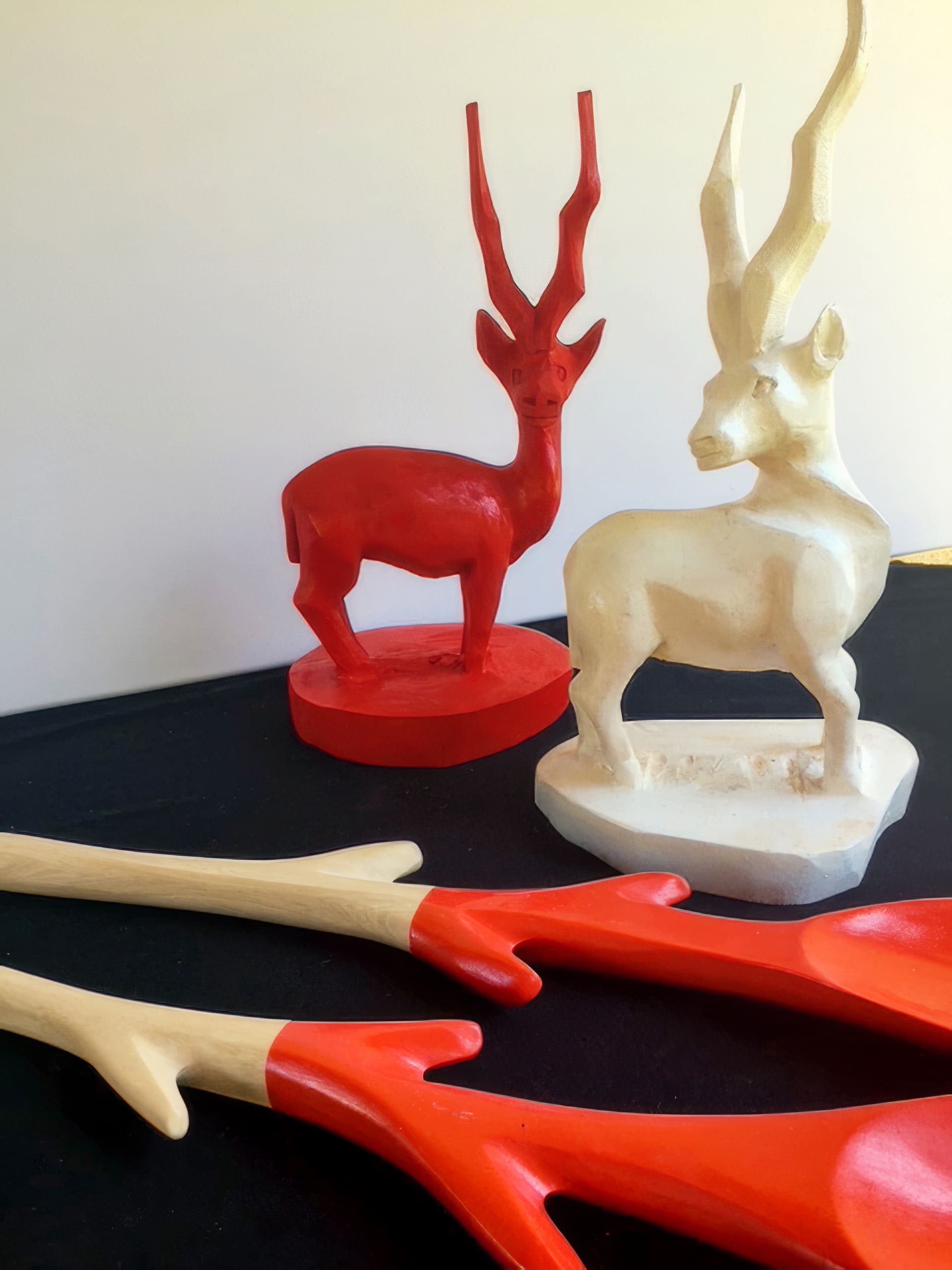Two wooden antelope sculptures—one painted red, one white—stand on a black surface against a plain background. In the foreground lie two wooden utensils with natural handles and red-painted pronged ends, mimicking the colouration of the red antelope.