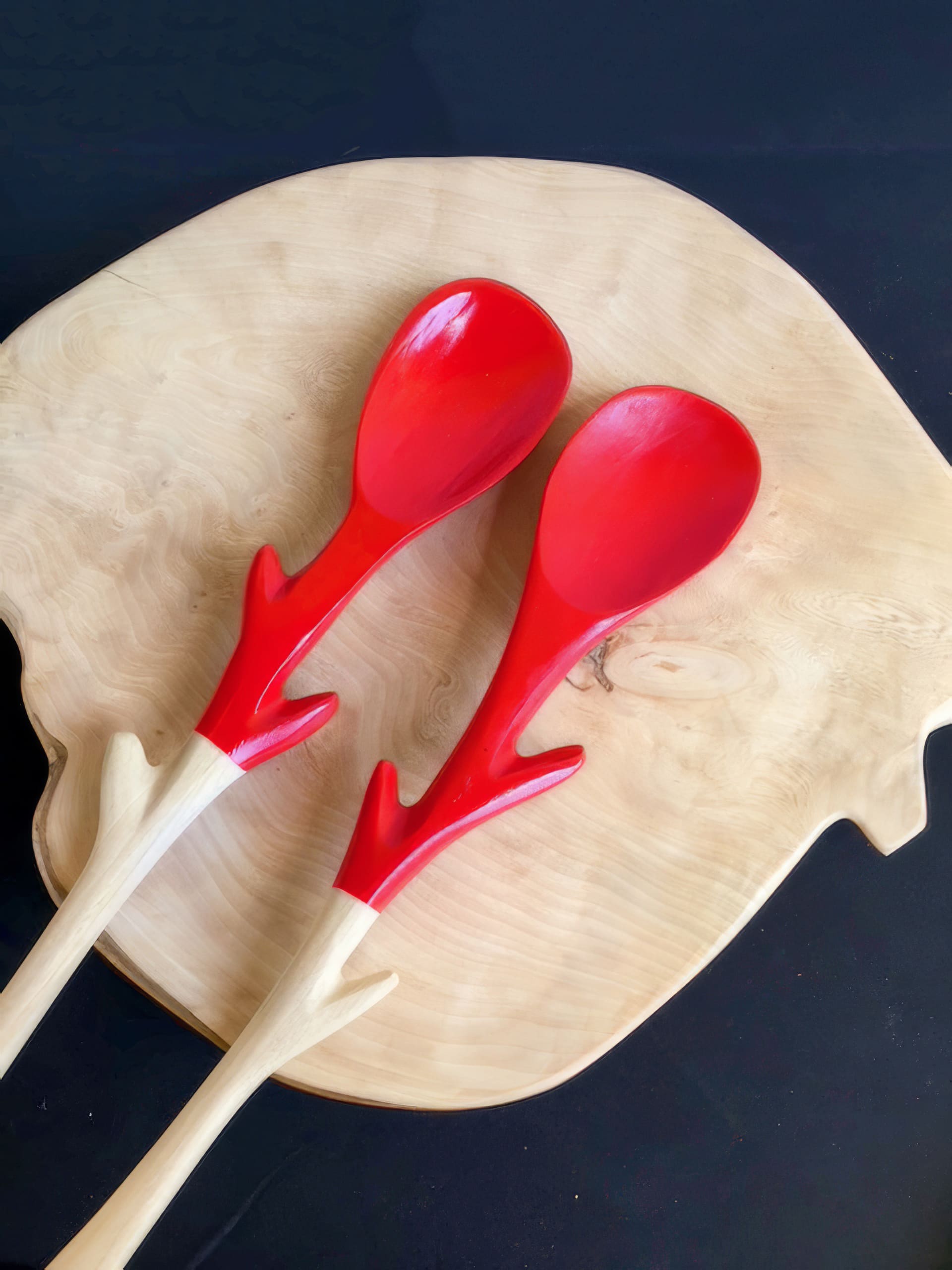 Two wooden spoons with red, antler-shaped handles rest on a light-coloured, irregularly shaped wooden chopping board, set against a dark background.