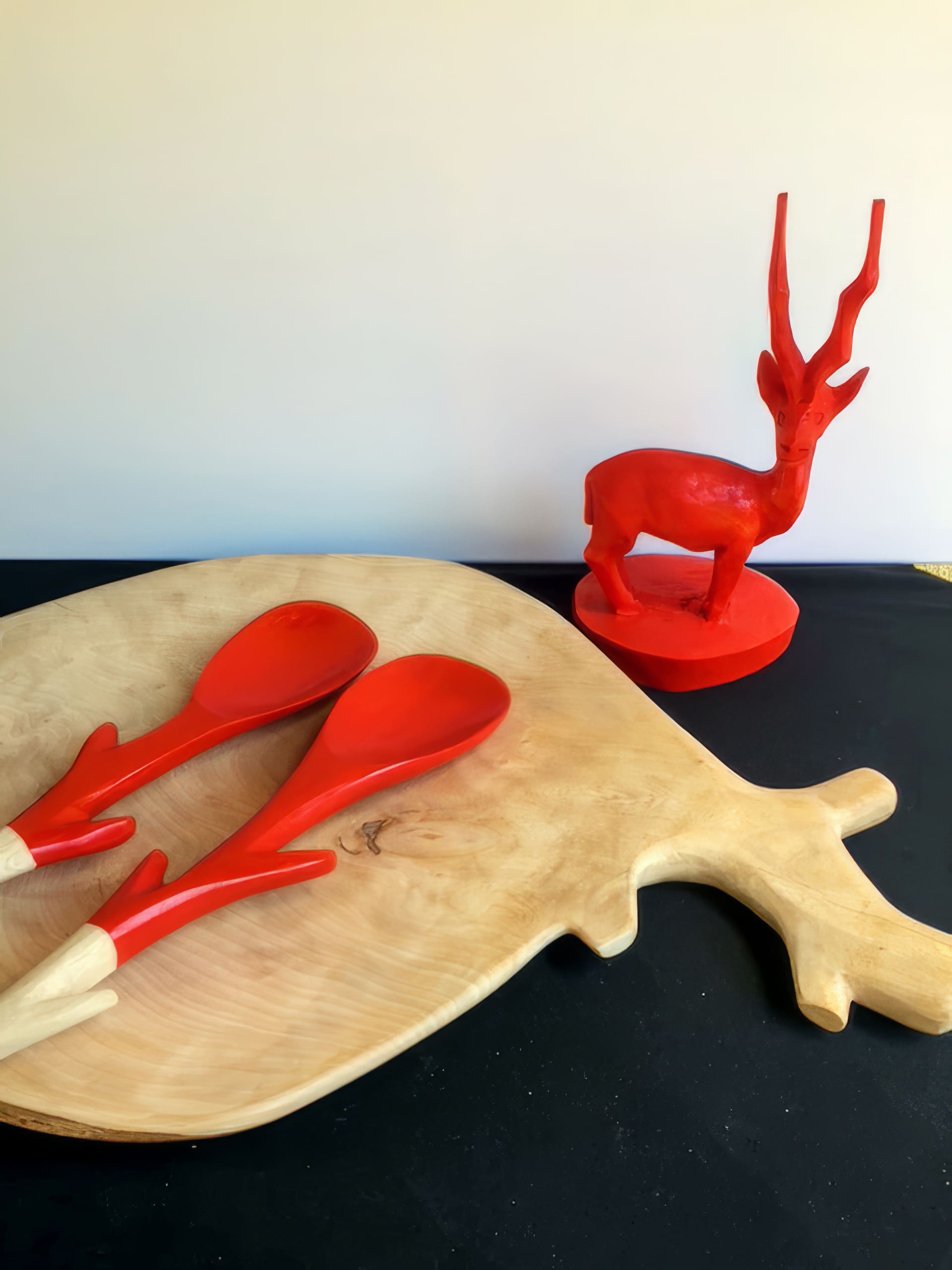A wooden chopping board shaped like an animal with antlers, two red serving spoons with antler-like handles, and a bright red antelope figurine are arranged on a black and light surface against a plain light background.