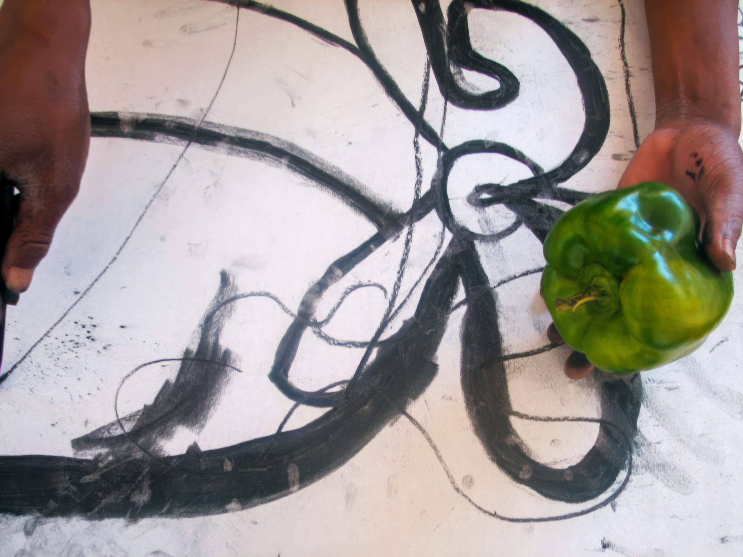 Two hands hold a black marker (left) and a green pepper (right) over a white surface with bold black abstract lines and swirls, suggesting the pepper may inspire the drawing’s shapes.