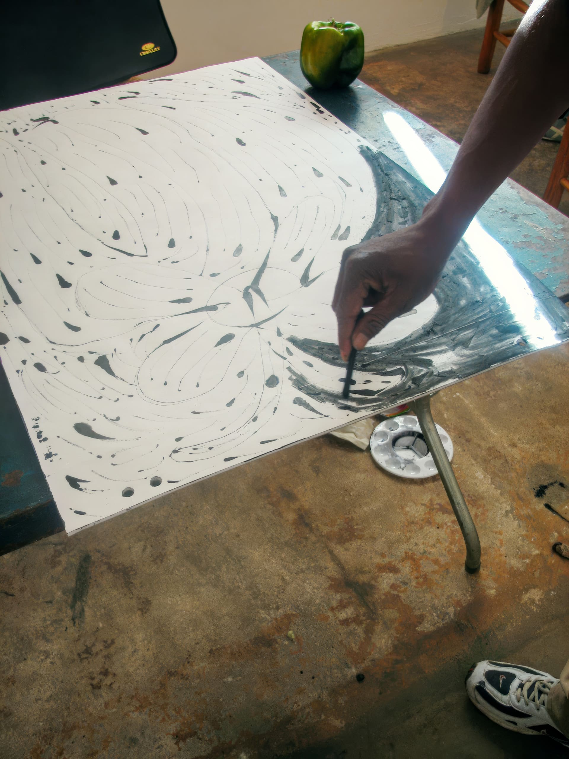 A person’s hand uses black paint and a brush to create swirling, abstract patterns on a large sheet of white paper. The artwork is on a table with paint supplies nearby. A green pepper sits on the table, and the floor is stained and textured.