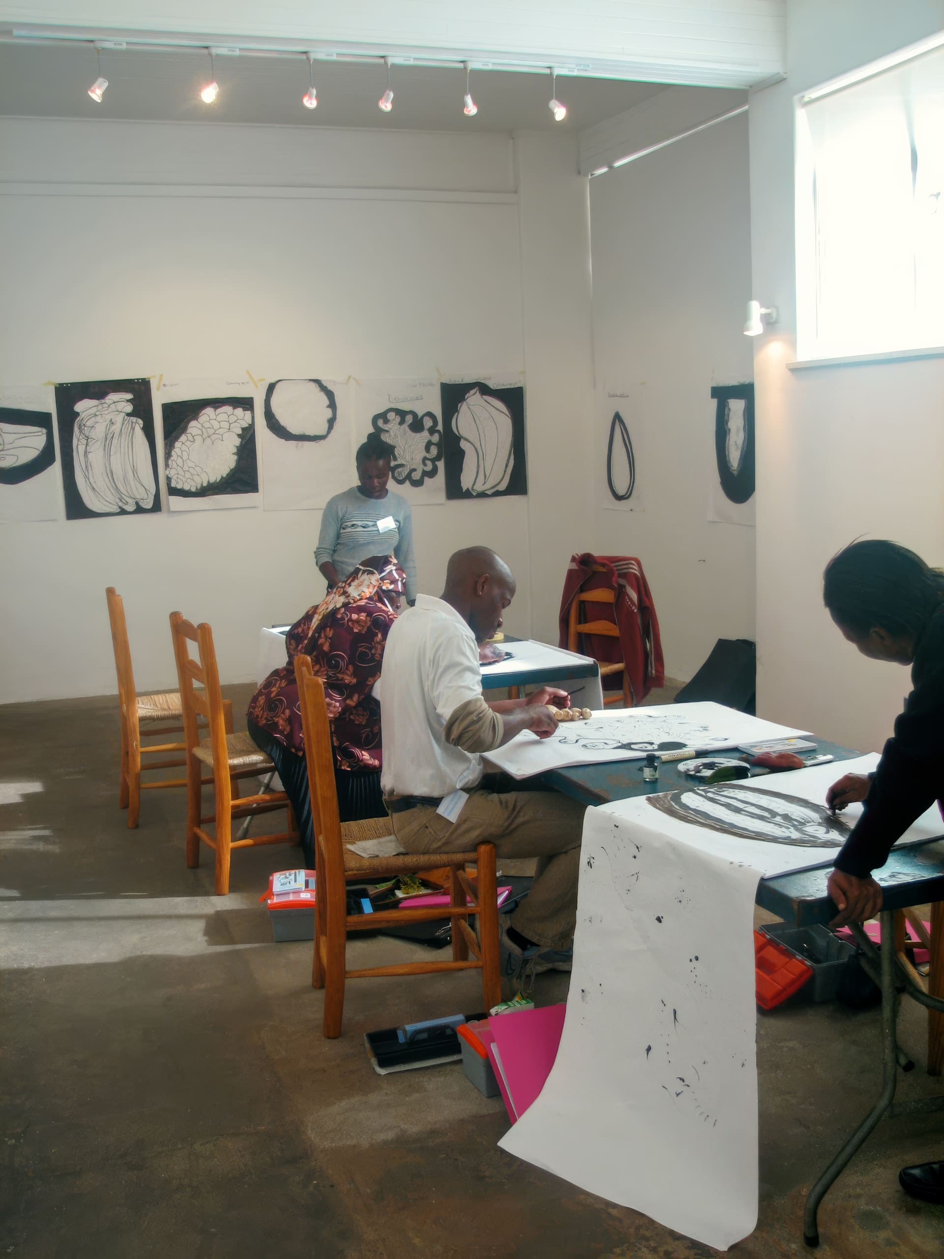 Three people sit at wooden tables in an art studio, sketching or painting on long sheets of paper. Black-and-white abstract art hangs on the walls. Another person stands in the background. The room is well-lit with natural and track lighting.