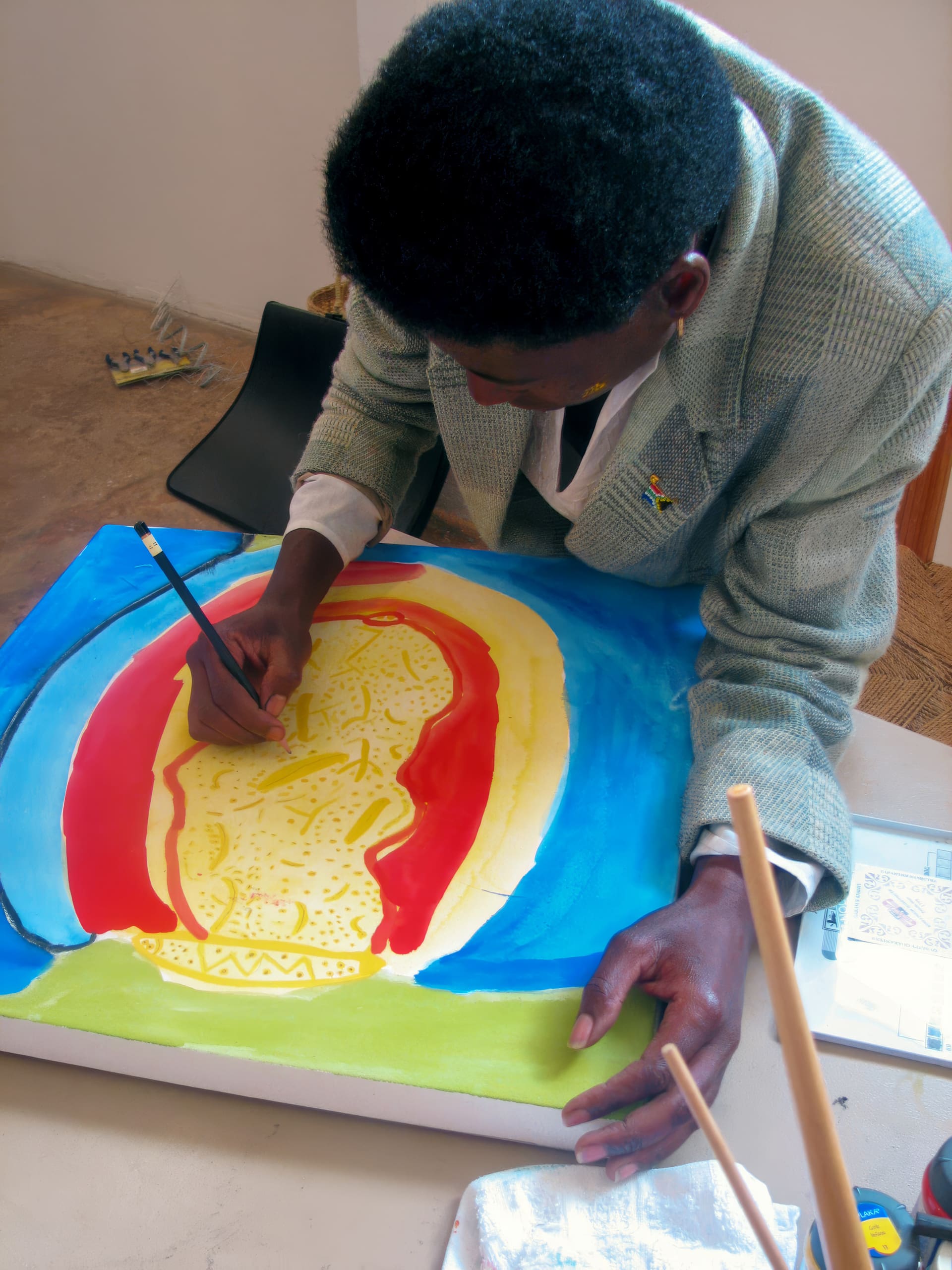 A person with short, curly hair and wearing a grey jacket is bending over a table, painting a colourful abstract image with yellow, red, and blue areas on a canvas. Art supplies and brushes are scattered on the tabletop in the foreground.