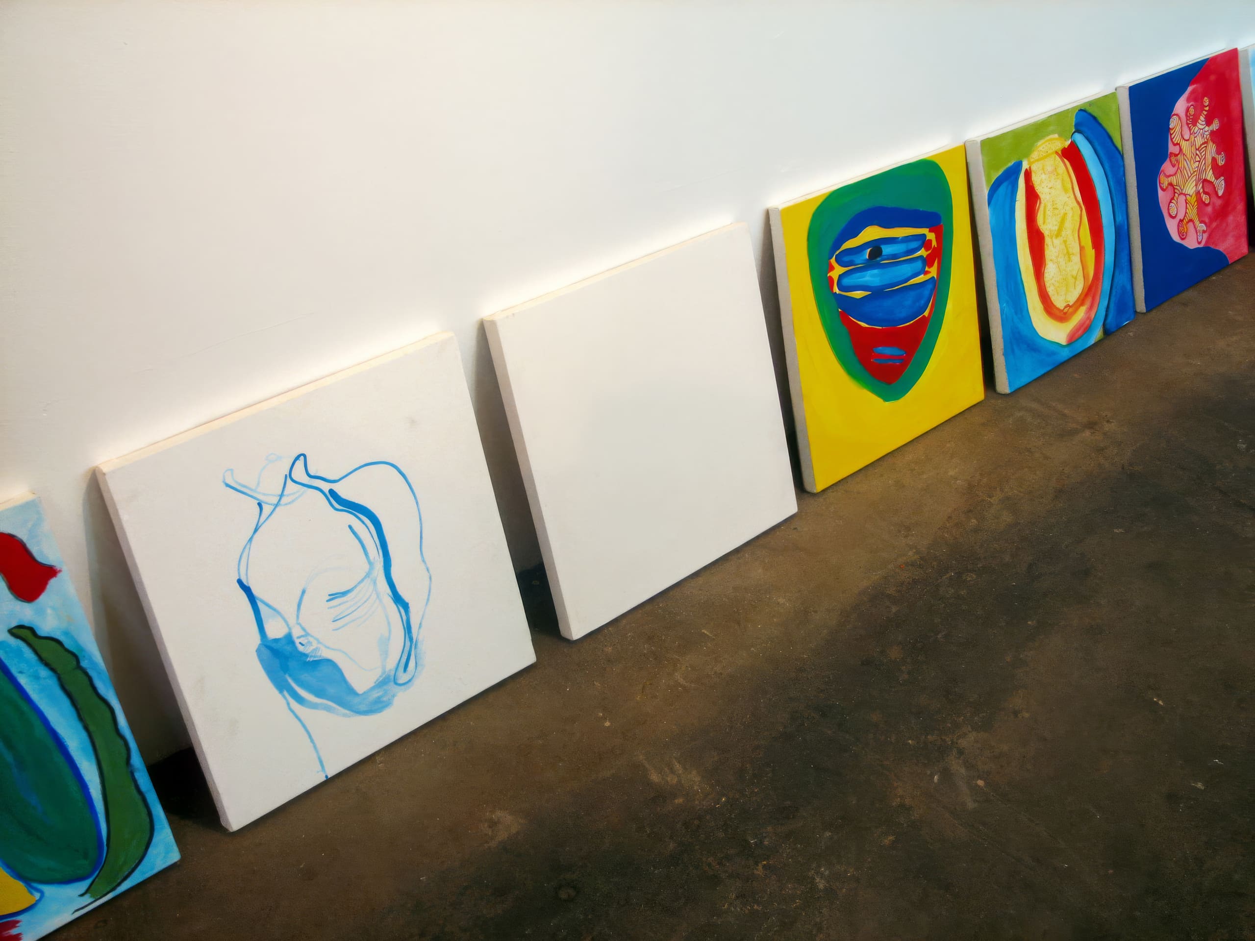 Six colourful abstract paintings lean against a white wall on a brown floor. Most feature bright colours and organic shapes; one canvas in the centre is blank. The artworks include blue, yellow, red, and green hues, each with a unique, expressive design.