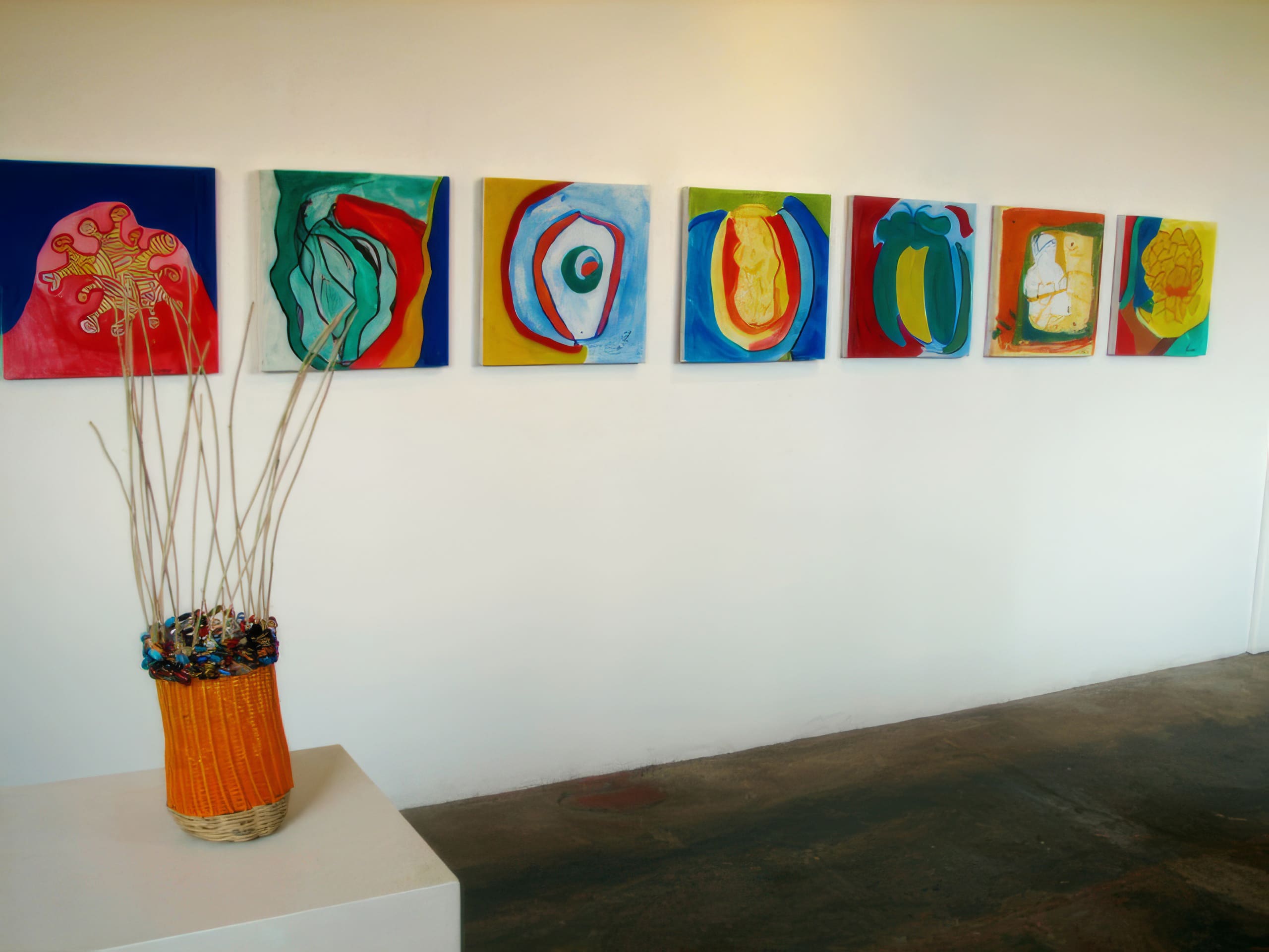 A row of eight colourful abstract paintings hangs on a white wall. Each canvas features bold shapes and vibrant colours. In the foreground, an orange basket with tall white sticks and multicoloured accents sits on a white pedestal. The floor is brown.