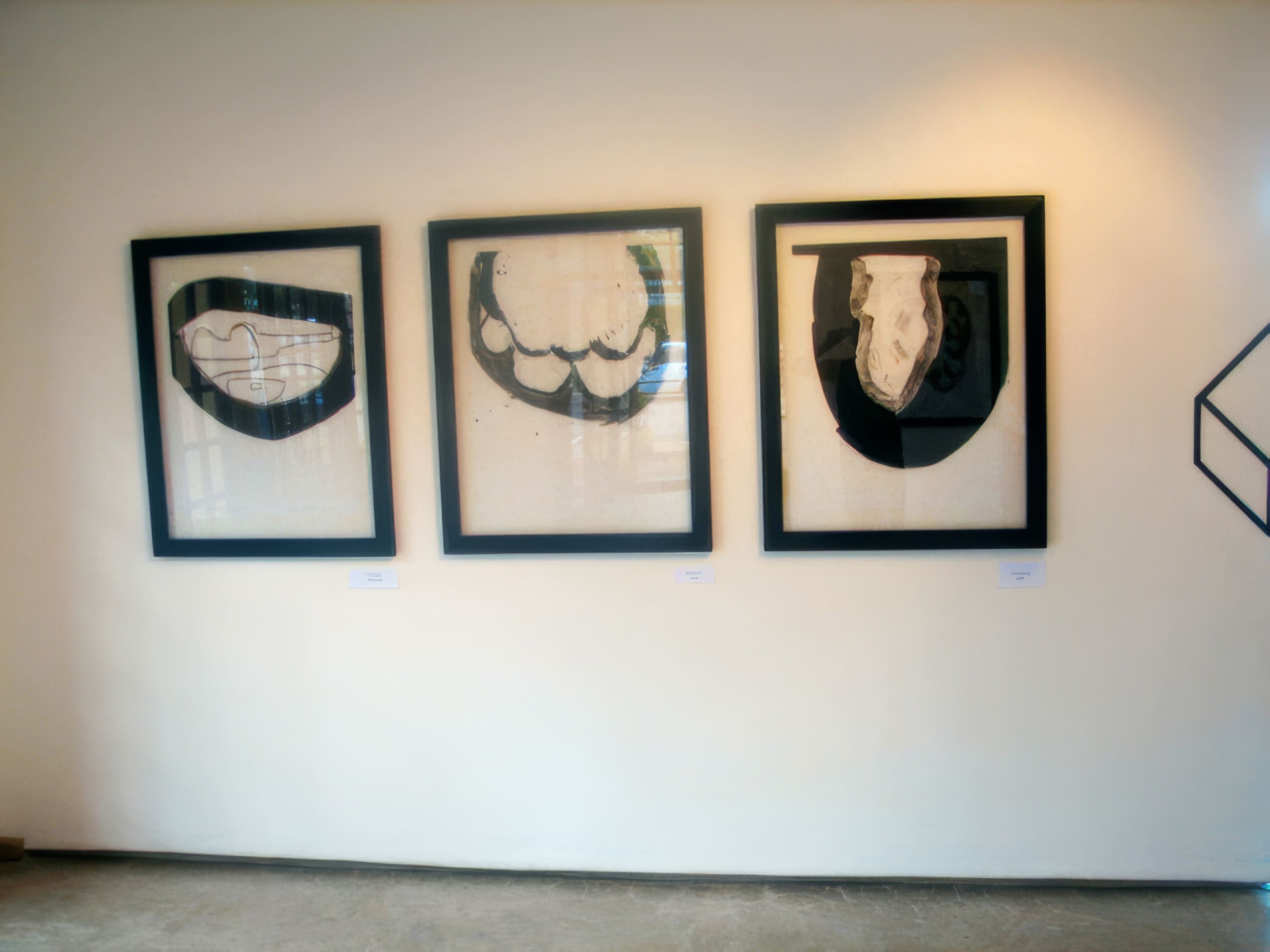 Three abstract black-and-white framed artworks hang on a plain cream-coloured wall. Each piece features bold, curved black shapes resembling stylised organic forms. Small white labels are beneath each frame. A partial geometric drawing appears on the right wall.