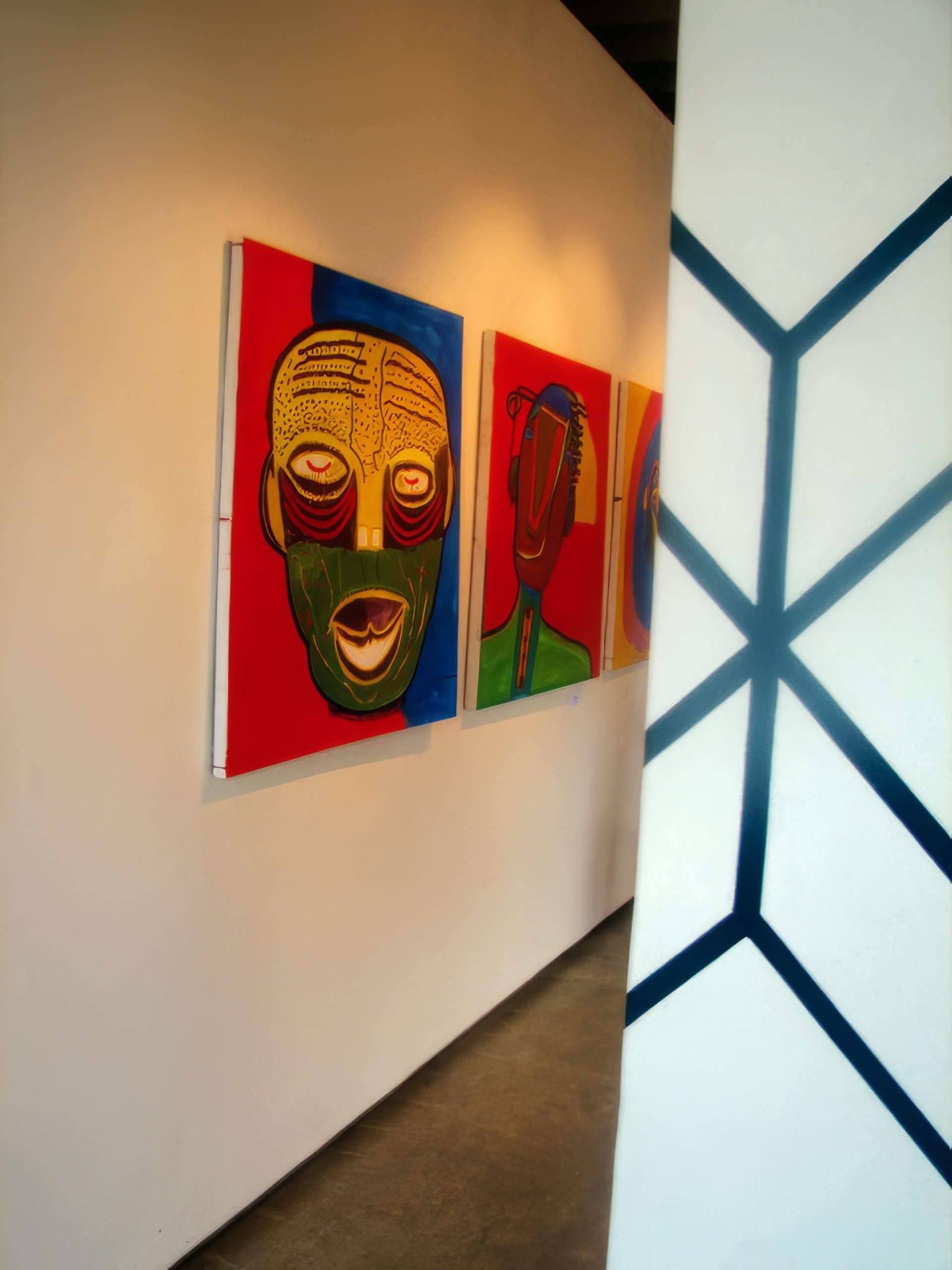 A modern art gallery displays colourful abstract portraits with bold lines and bright backgrounds on a white wall. In the foreground, a wall with a black geometric pattern partially obscures the view. The floor is polished concrete.