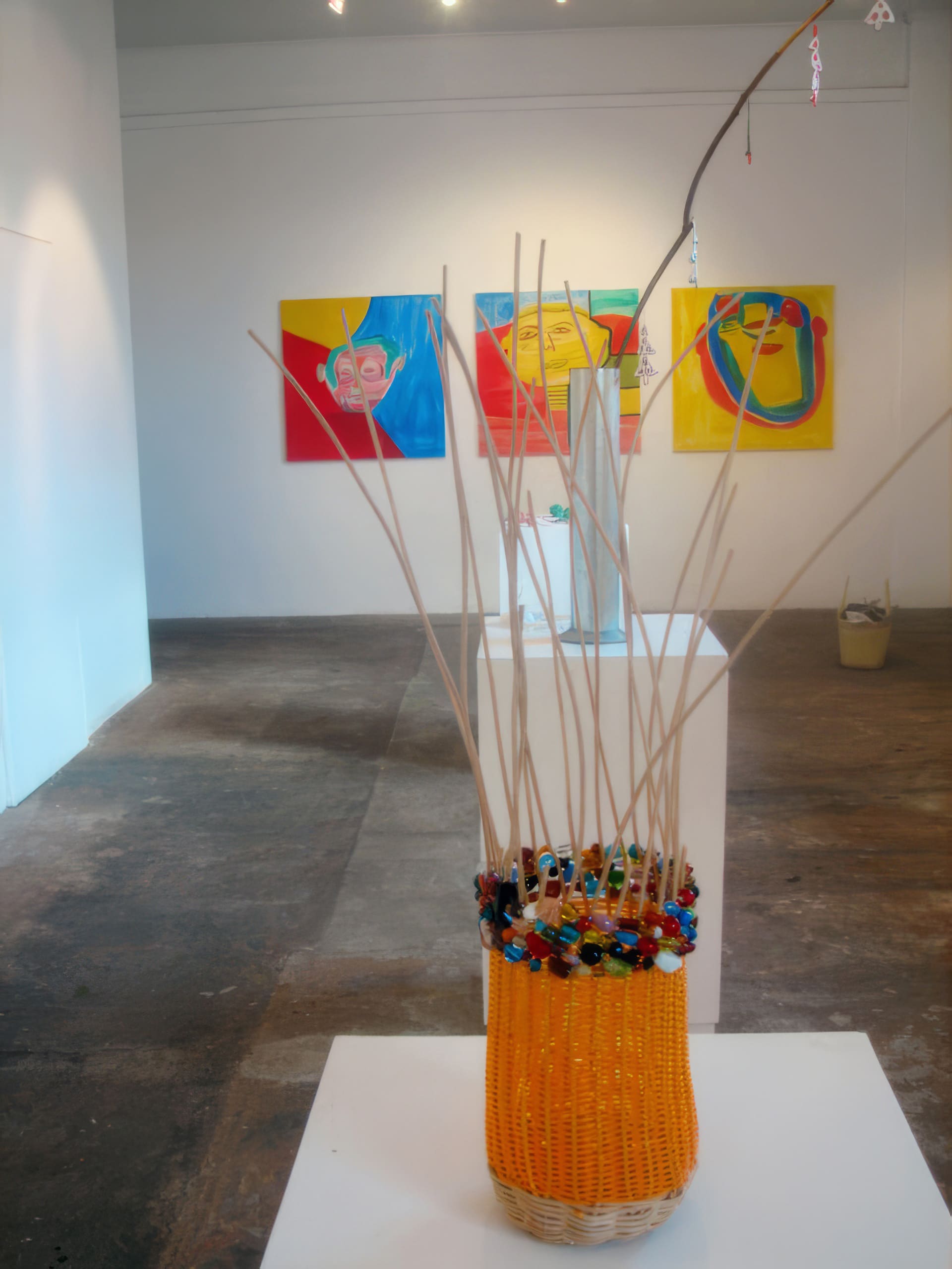 A bright yellow woven basket adorned with colourful beads and tall wooden sticks sits on a white plinth in a gallery. In the background, three vivid abstract paintings hang on a white wall, and another sculpture is visible further inside the space.
