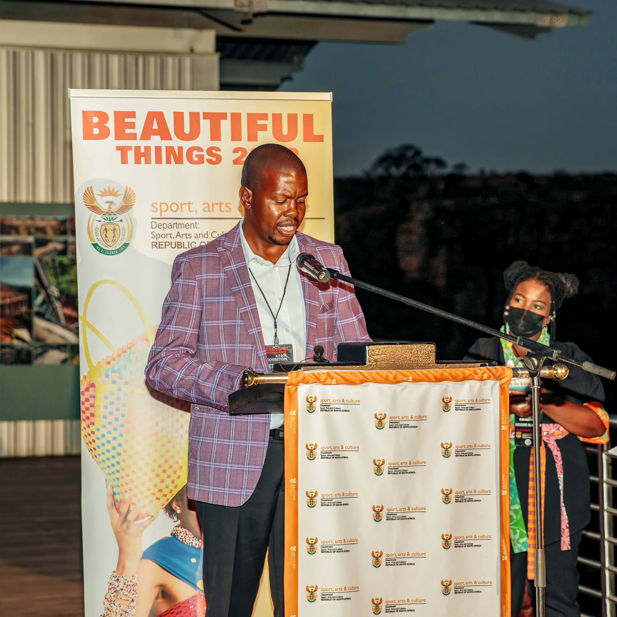 A man in a purple checked blazer speaks at a lectern with a microphone. Behind him is a banner reading “BEAUTIFUL THINGS 2.0” and a woman wearing a face mask stands to the side. It appears to be an official event related to sport, arts, and culture.