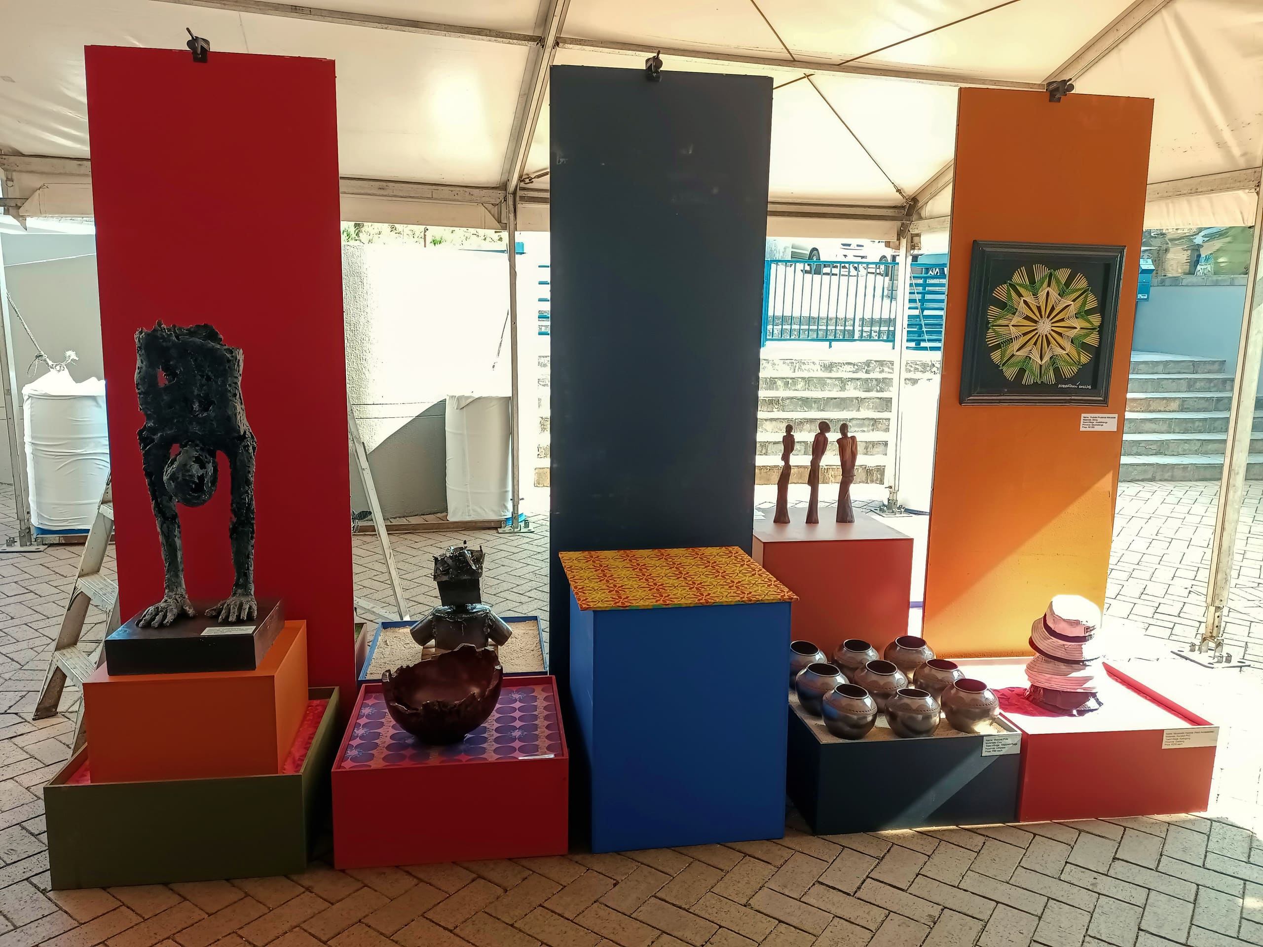 A display of various sculptures and pottery is arranged on colourful plinths under a marquee. The setup features abstract figures, vases, a black crouching figure, wooden statuettes, bowls, and a framed circular geometric artwork on bright orange and blue panels.