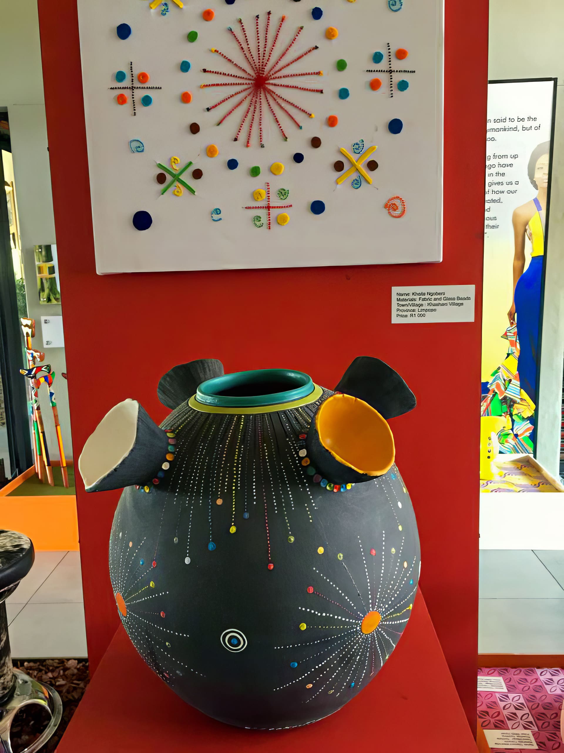 A large round black vase with four wide, colourful spouts and dotted, starburst patterns sits on display. Above it, a white canvas features geometric, starburst designs made from colourful beads and string, mounted on a red wall in a vibrant art exhibit.
