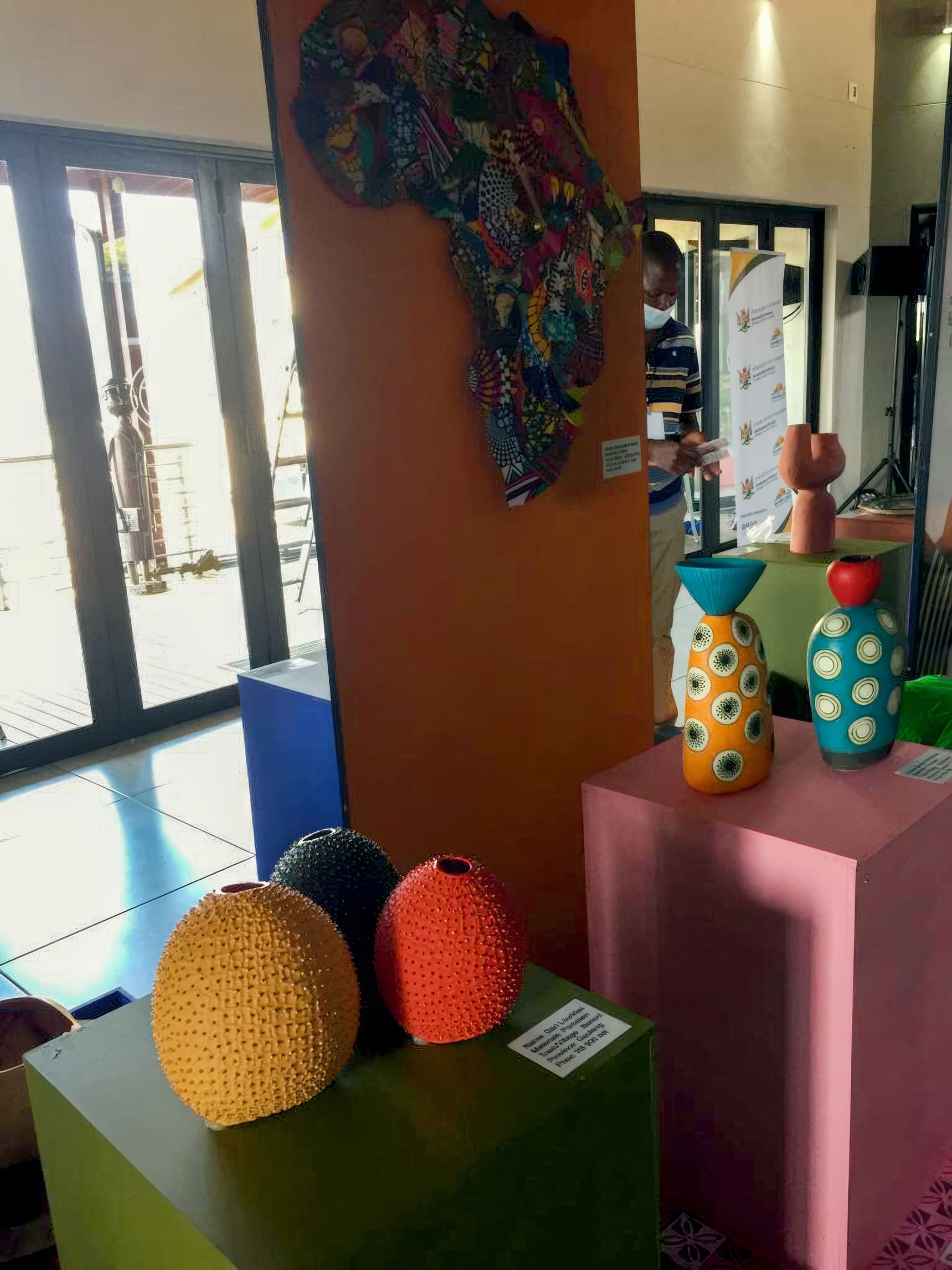 Colourful ceramic vases with textured surfaces are displayed on blue and pink plinths. Behind them is a vertical panel with a vibrant, patchwork Africa-shaped wall art. A person stands near large windows in the background.