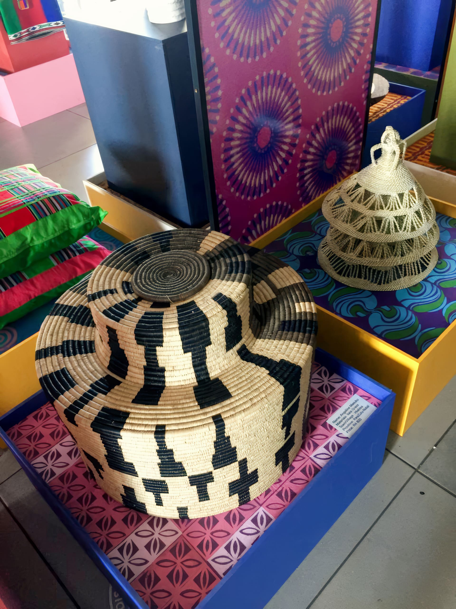 Colourful display of woven hats and textiles. Foreground: large, round, straw hat with bold black geometric patterns. Behind it is a conical, tan hat with intricate weave. Background items include patterned cushions and panels with vibrant circular designs.
