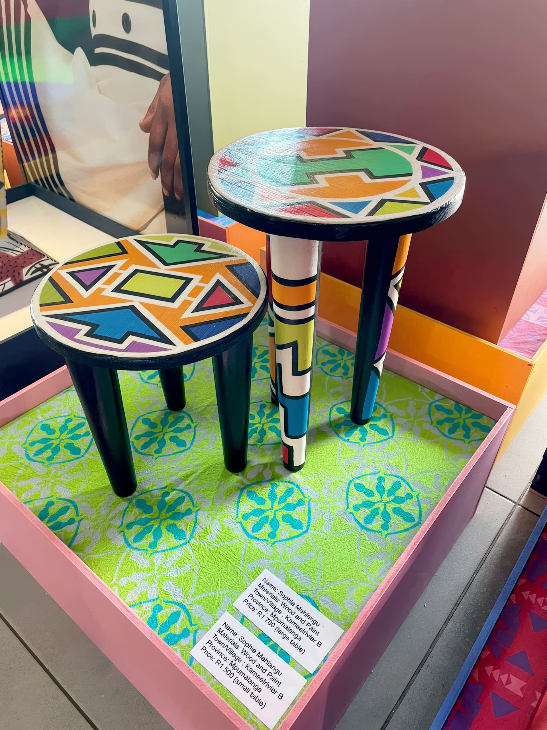 Two round stools with colourful, geometric patterns on the seats and legs sit on a patterned green platform in a display case. A background image shows a person, and labels with details about the stools are visible on the display’s edge.